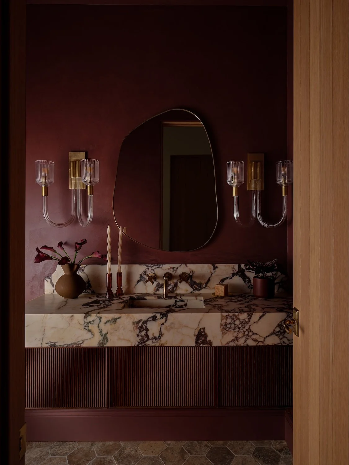 A bathroom with a marble Moody burgundy powder room with dramatic marble vanity, fluted wood cabinetry and sculptural mirror, a mirror, and wall-mounted light fixtures on either side of the mirror.