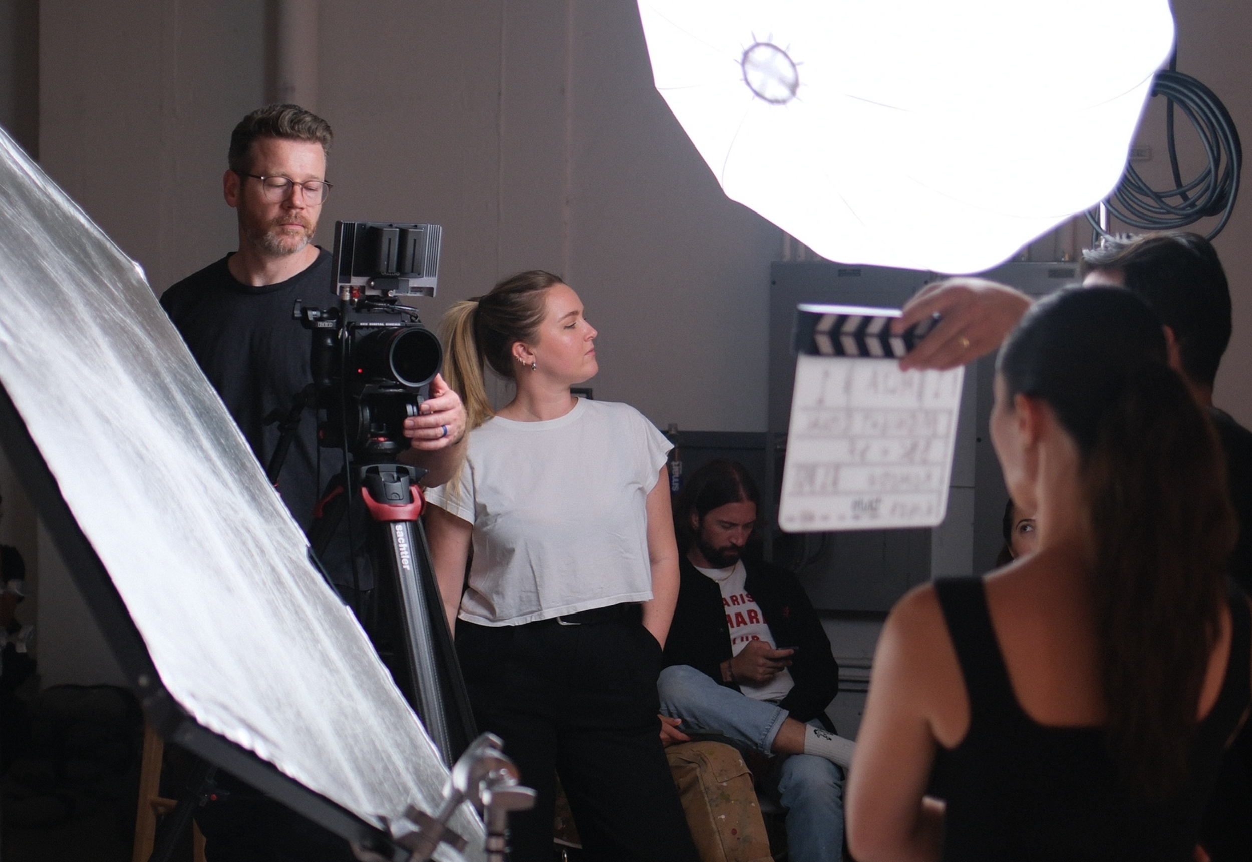 Film crew on set preparing for a scene, Camera operator Stephen Alberts films with a woman standing among equipment, a Director, and lighting gear visible.