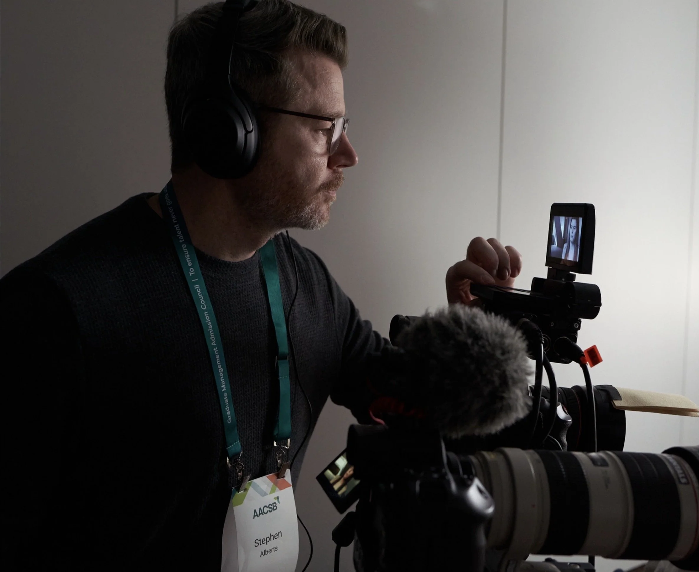 Stephen Alberts headphones and glasses looks through a camera with a mounted monitor, capturing a scene of a woman on the screen. He has a badge and is wearing a lanyard for an event or conference.