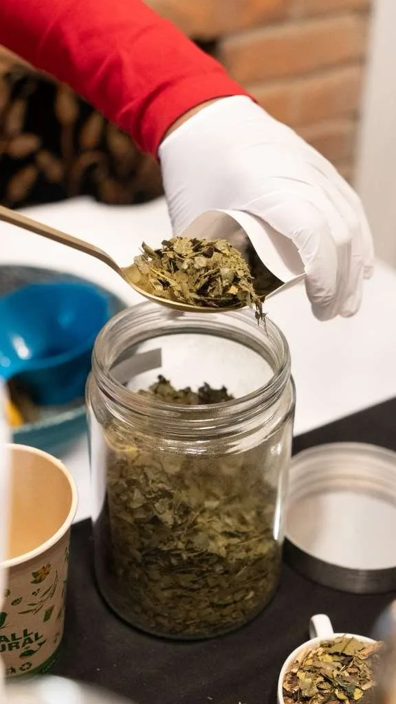 Person in a white glove and red sleeve scooping dried herbs from a glass jar with a spoon. There are other containers and a book nearby.