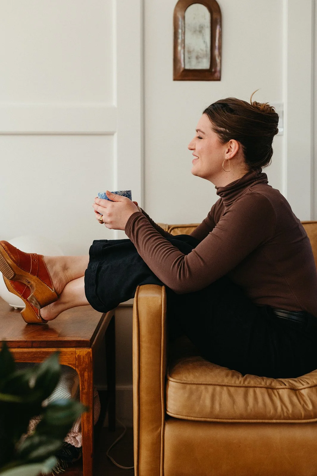 A woman with brown hair tied up, sitting on a brown couch with her legs resting on a wooden table, holding a blue mug, smiling, wearing a brown turtleneck and gold hoop earrings.