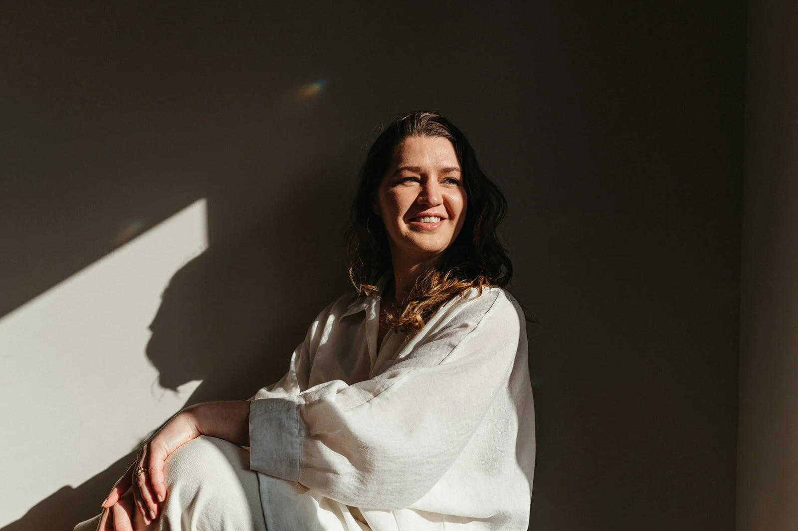 A woman sitting against a wall, smiling, with sunlight casting shadows on her face and wall behind her. She has shoulder-length dark hair and is wearing a white shirt.