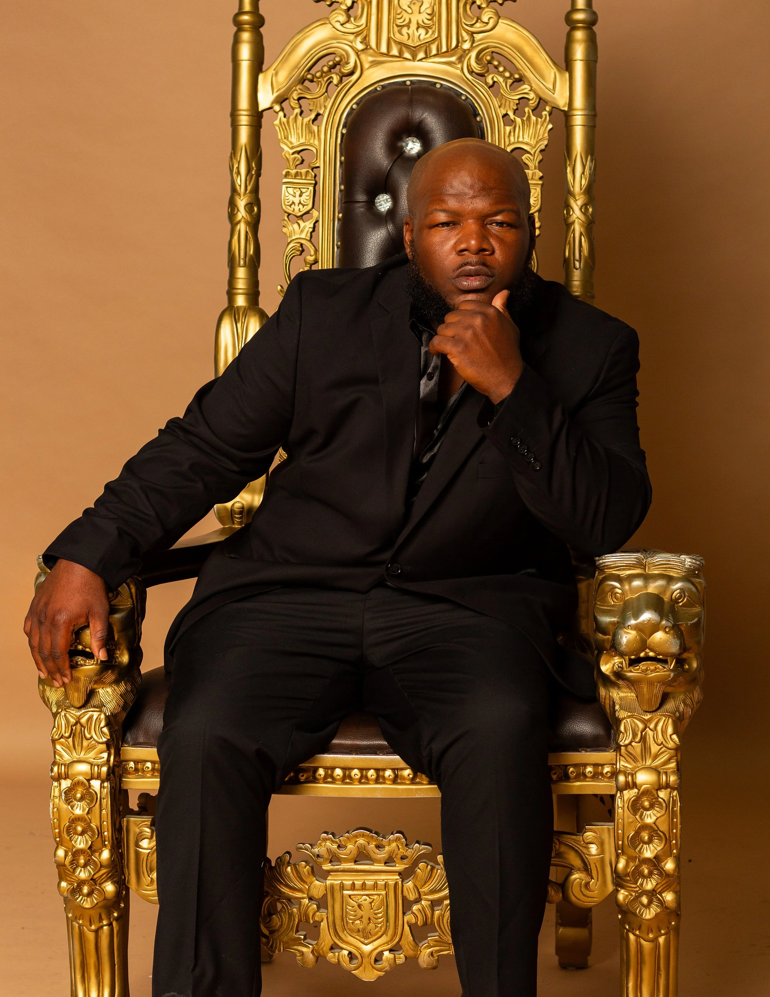A man sitting on an ornate, gold-colored throne with intricate carvings, wearing a black suit and resting his chin on his hand, looking serious.