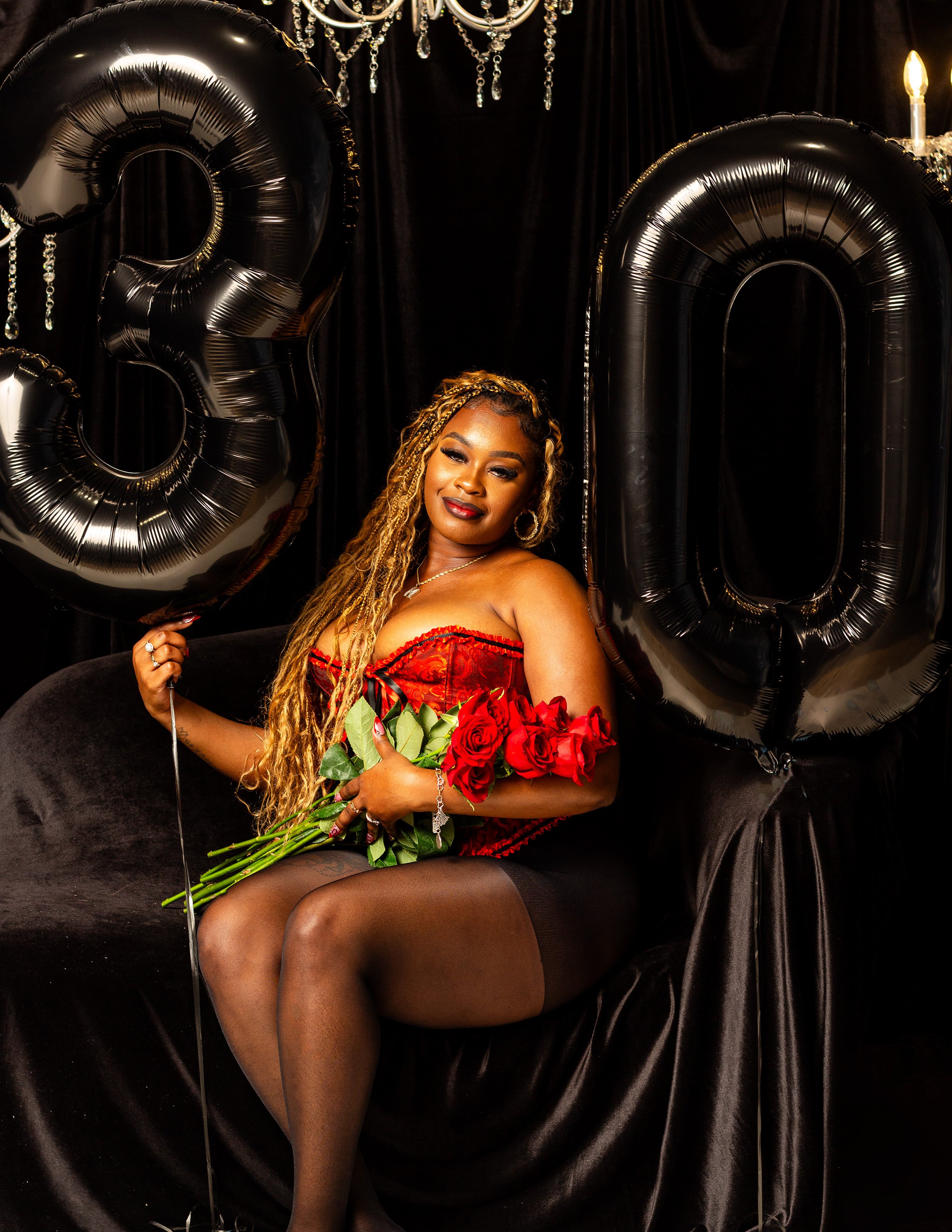 Woman with long blonde braided hair and red lipstick sits on a black velvet chair, holding a bouquet of red roses in her arms, surrounded by large black balloons shaped as the numbers 2 and 0, with a black curtain backdrop and hanging crystal decorations.