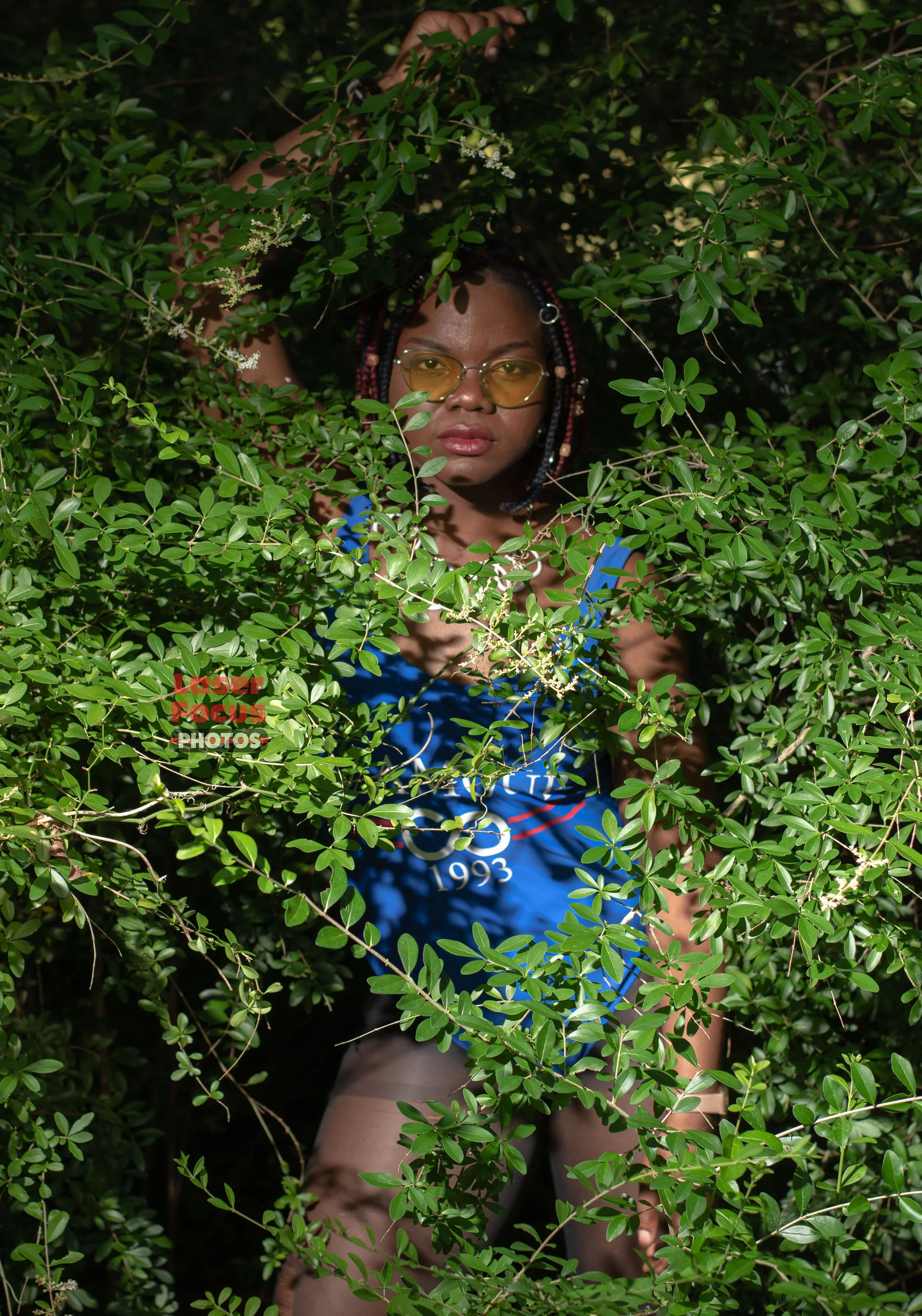 A woman with glasses and braided hair partially hidden by green leafy bushes, wearing a blue tank top.