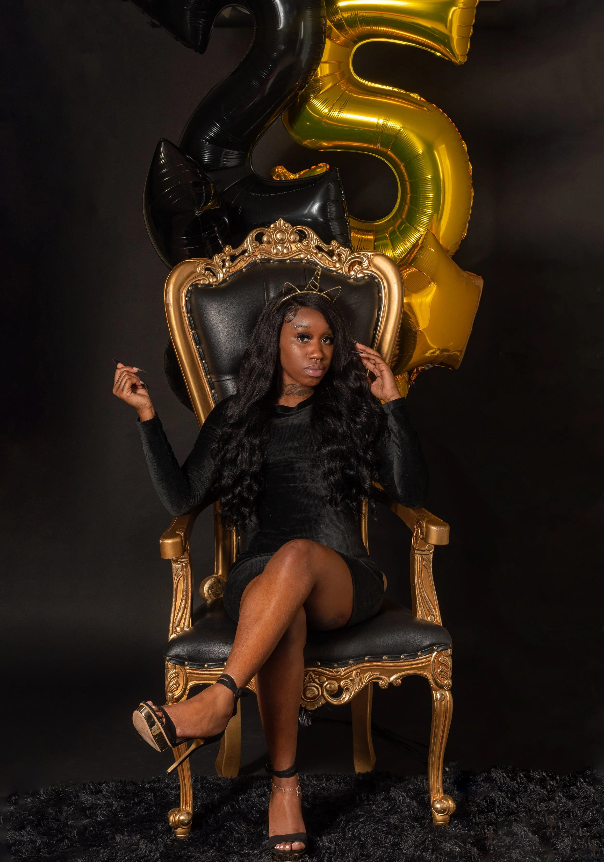 A woman with long wavy black hair sitting on a black and gold ornate throne chair against a black background. She is wearing a black dress, high heels, and has a gold headband with a small horn on her head. Behind her, there are large gold and black 