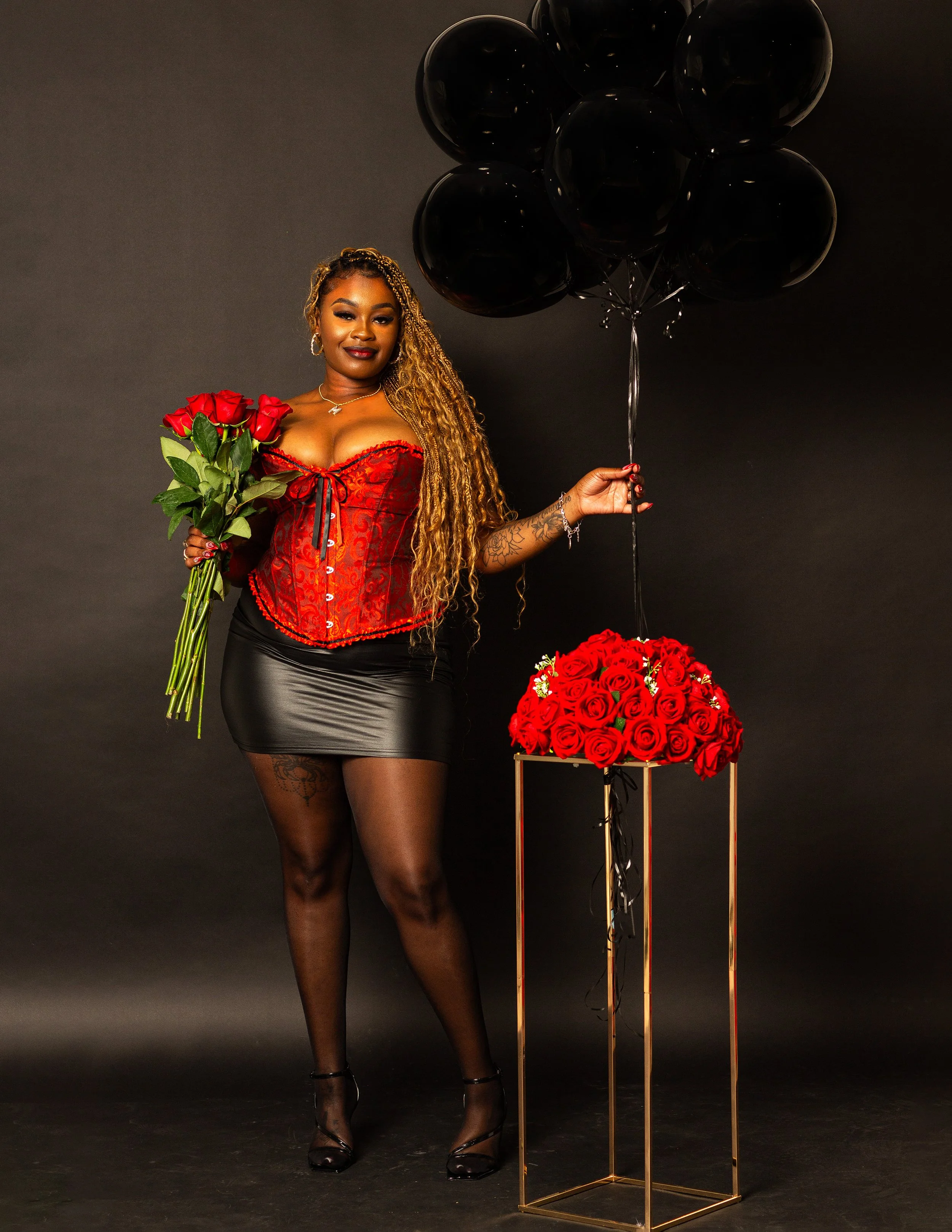 A woman with long, curly, blonde hair wearing a red corset top, black leather skirt, and black tights, holding a bouquet of red roses in her left hand and black balloons in her right hand, standing against a dark background.