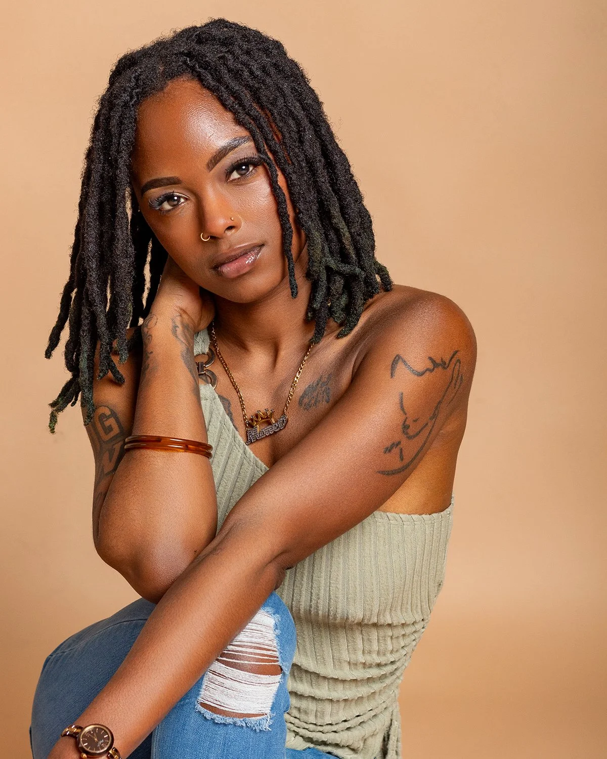 A woman with dreadlocks poses with her hand near her face, wearing a tank top, ripped jeans, and jewelry, against a beige backdrop.
