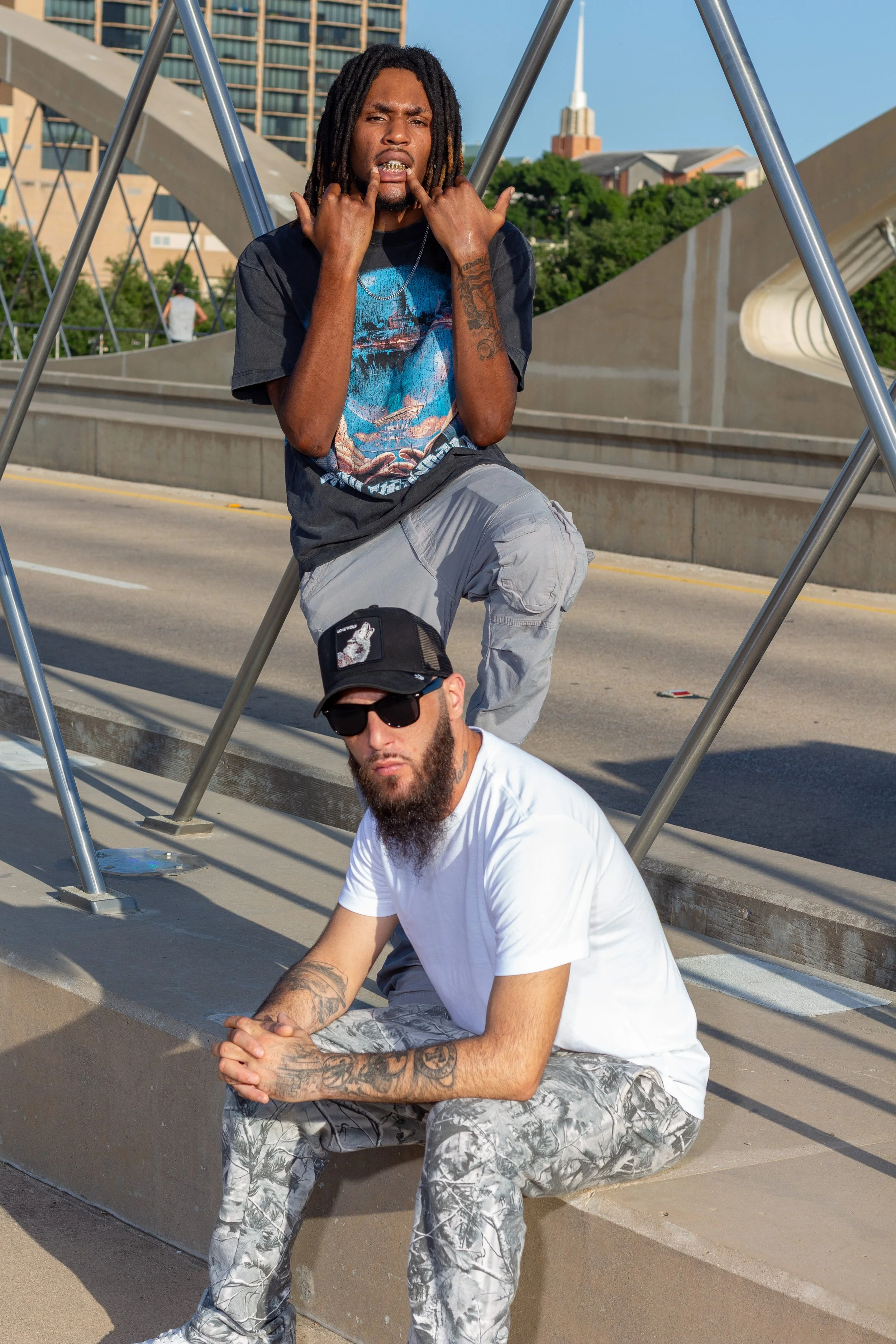 Two men sitting on a concrete ledge outdoors on a sunny day with an urban background. The man in the foreground has a beard, sunglasses, a black cap, and is wearing a white T-shirt with rolled-up sleeves and camouflage pants. The man in the backgroun
