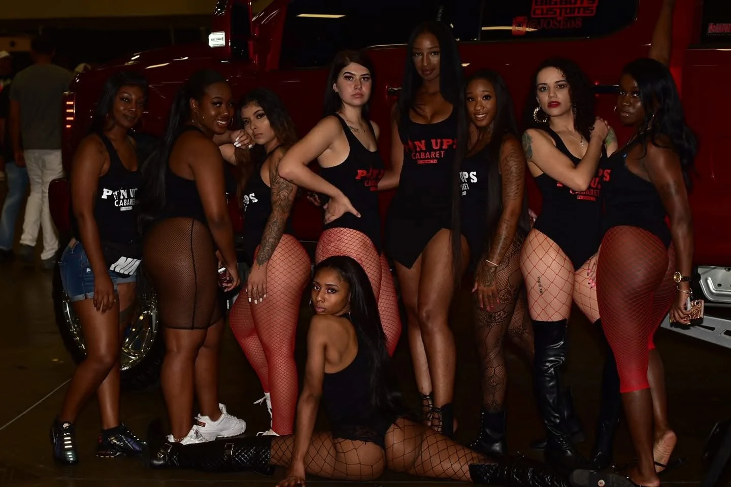 Group of nine women posing together in front of a red vehicle in a dimly lit indoor setting, wearing black tops and fishnet stockings.
