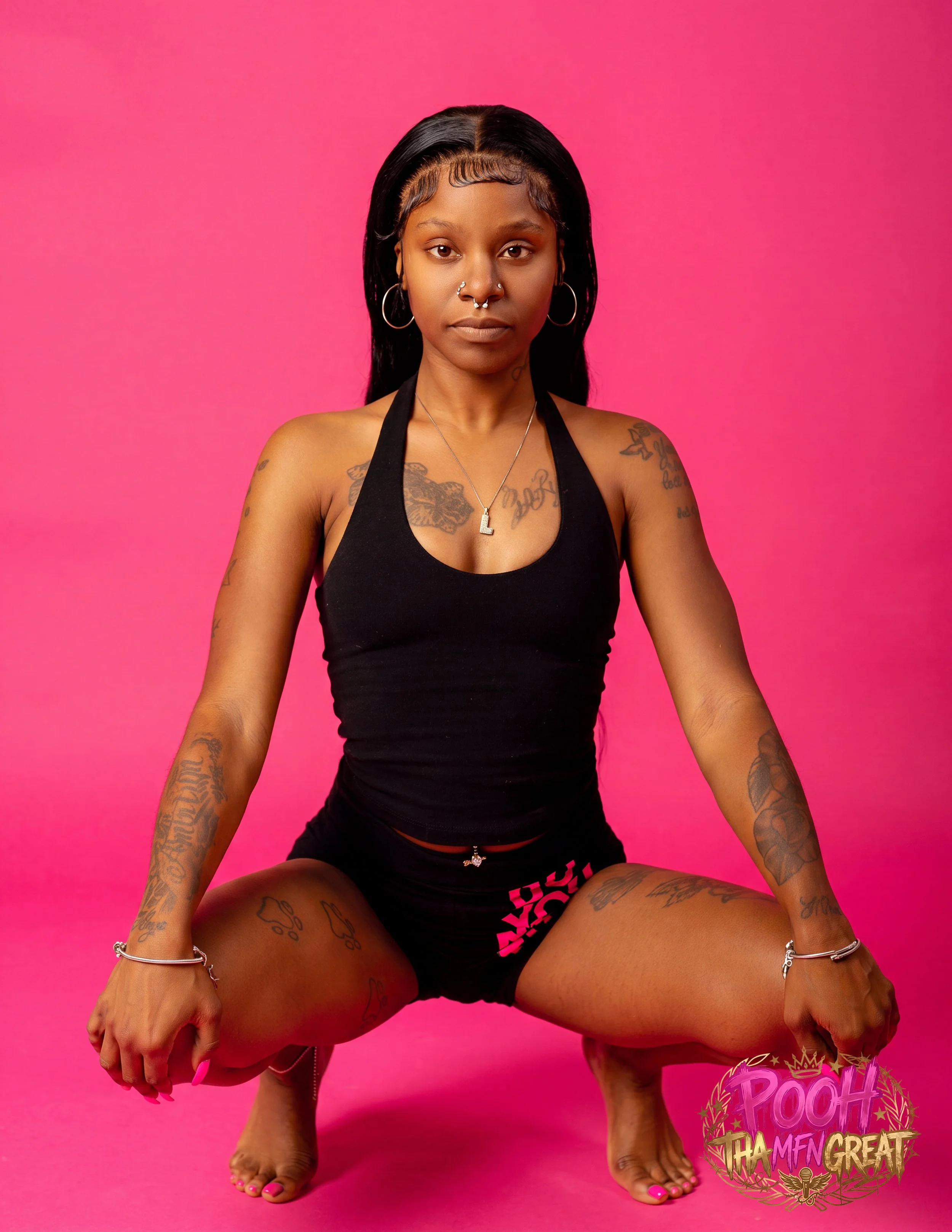 Young woman with black hair and tattoos, sitting against a pink background, wearing a black sports bra and shorts, with a serious expression.