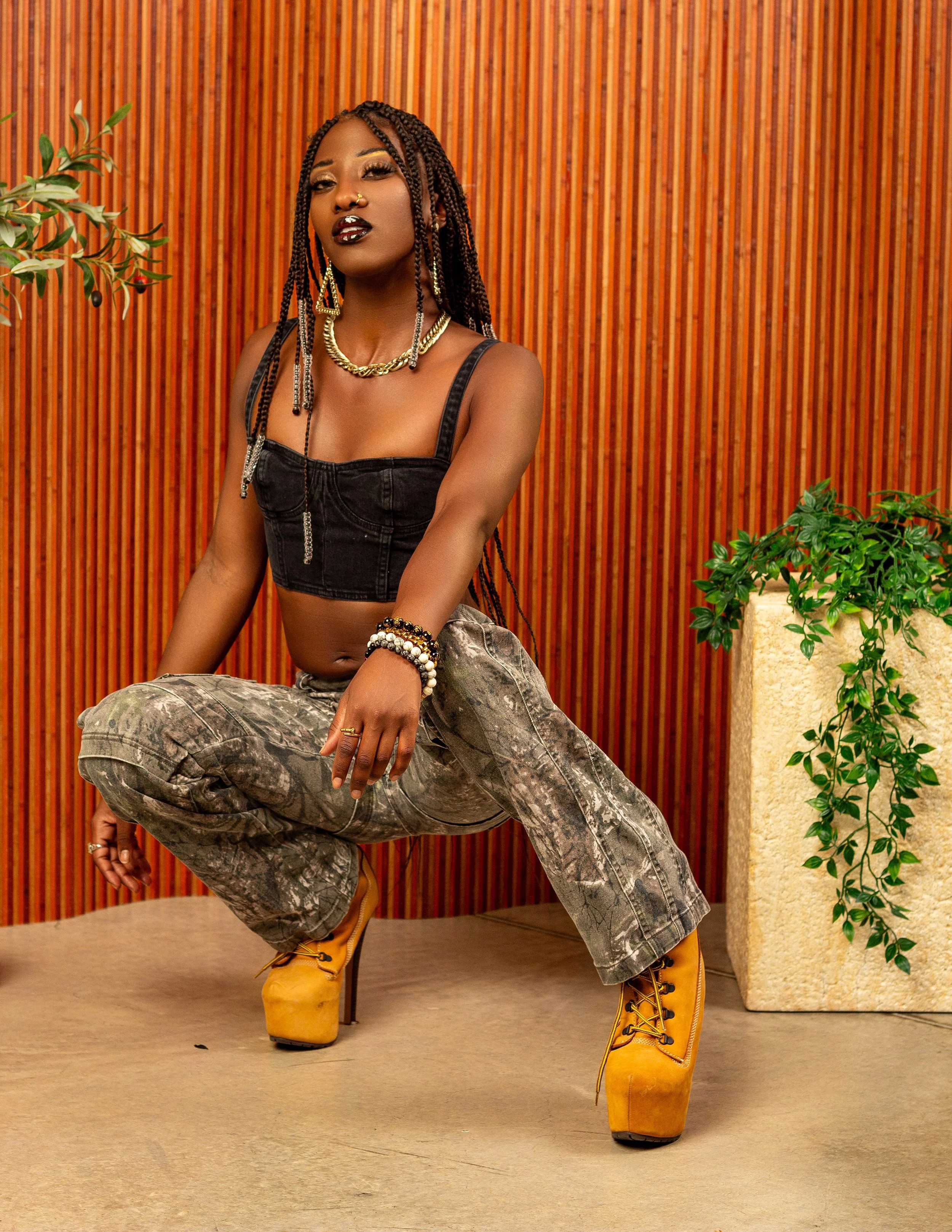A woman posing indoors with one leg bent and resting on a stool, wearing a black crop top, camouflage pants, yellow boots, and gold jewelry, with a striped wooden wall and green potted plant in the background.