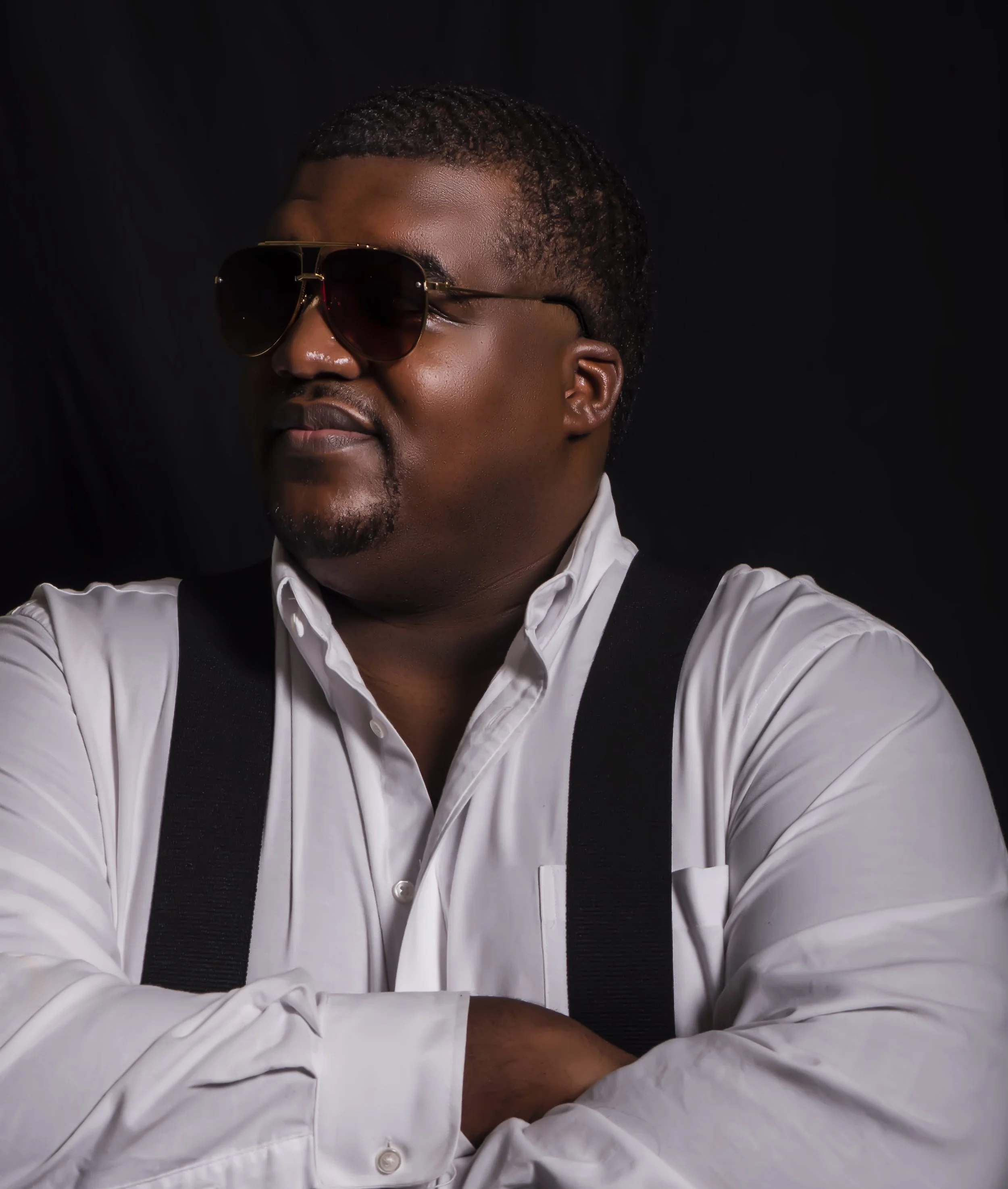 A man with dark skin wearing sunglasses, a white shirt, and a black vest on a black background.