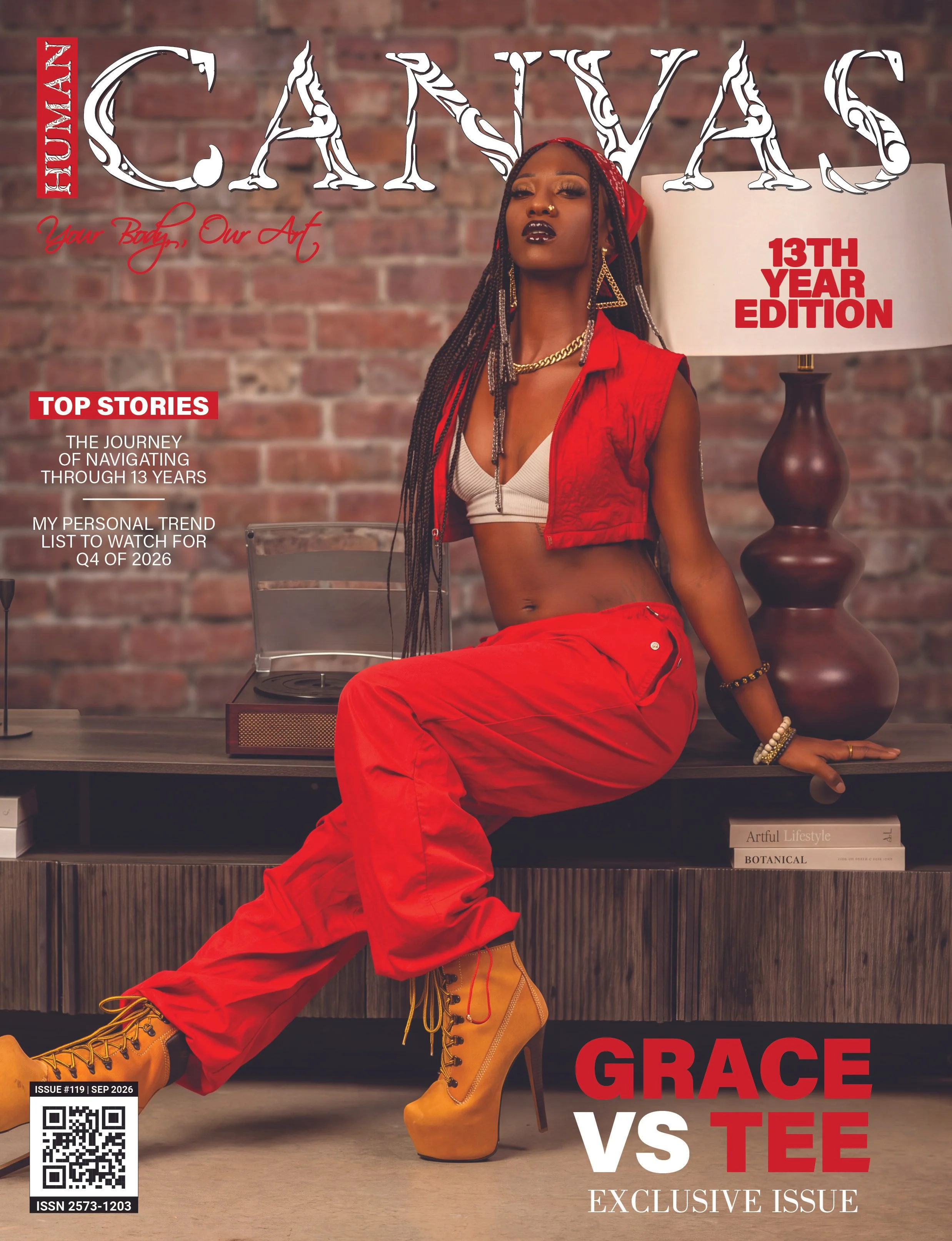 A woman with dramatic makeup and long braids wearing a red vest, white crop top, red pants, and tan combat boots posed in front of a brick wall and a table with books, along with a lamp indicating a magazine cover.