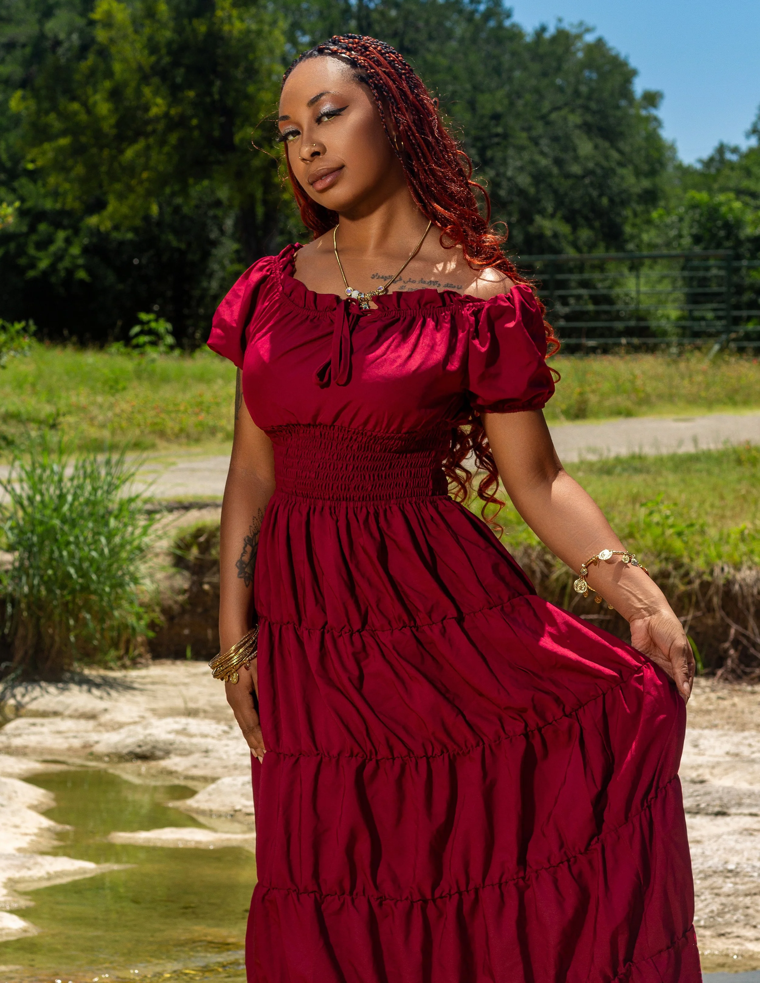 A woman with braided red hair standing outdoors near a small pond in a park, wearing a dark pink dress and gold jewelry.