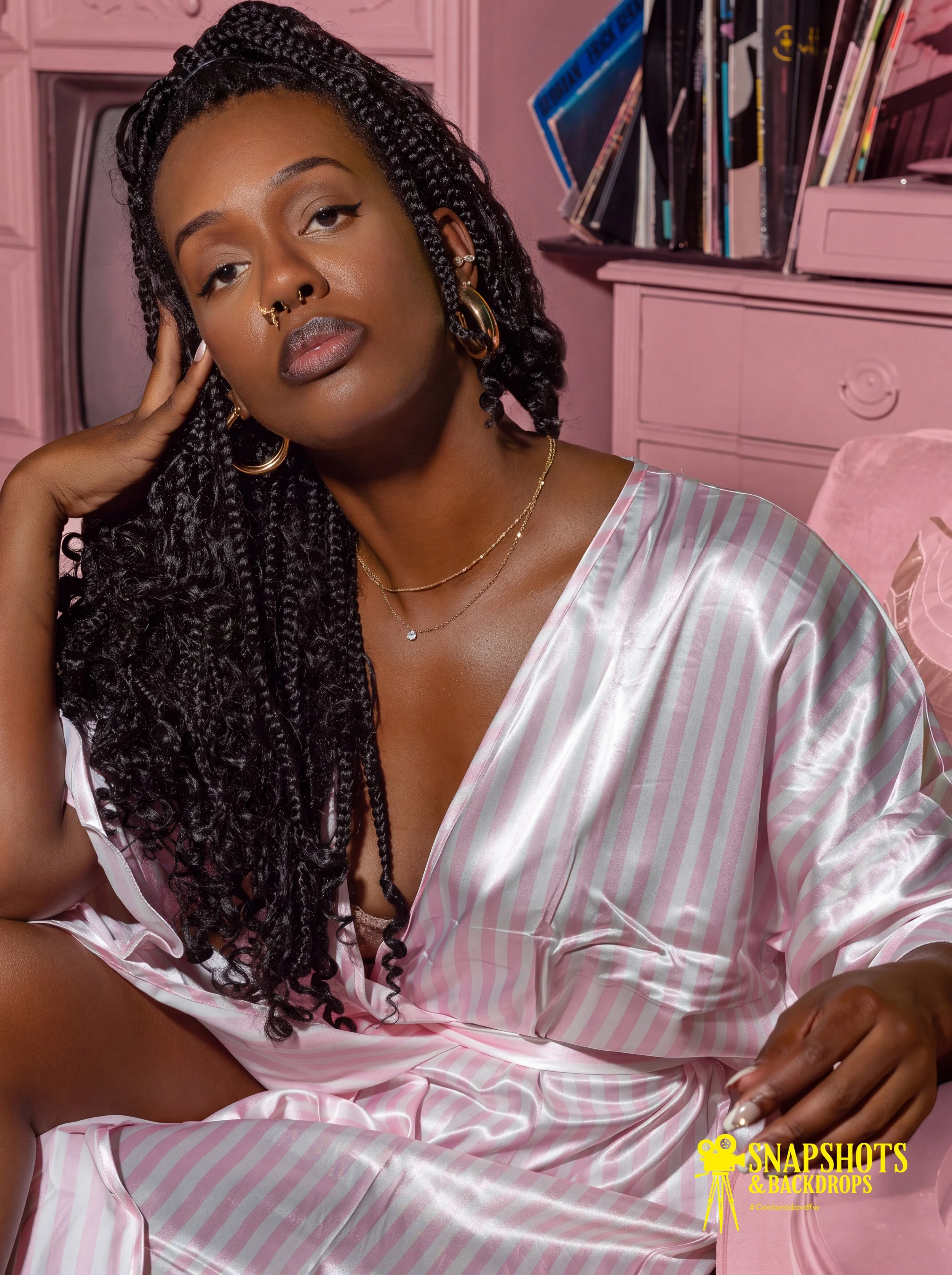 A woman with dark skin and long curly black hair styled in braids, resting her hand on her face. She has gold hoops, a septum piercing, and layered necklaces. She is wearing a pink and white striped satin robe, sitting in a pink room with a bookshelf