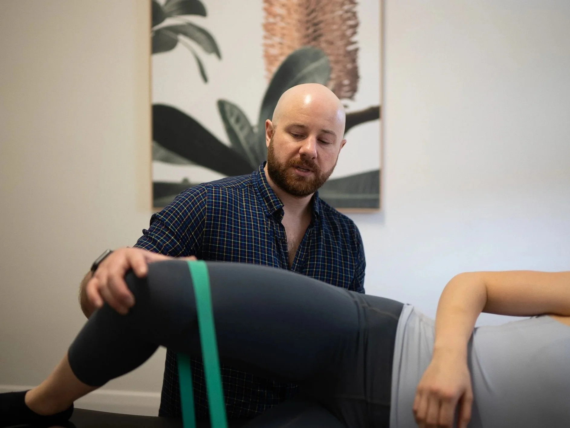 A physical therapist performing a stretching or therapy exercise on a patient lying on a treatment table, with a botanical print on the wall in the background.