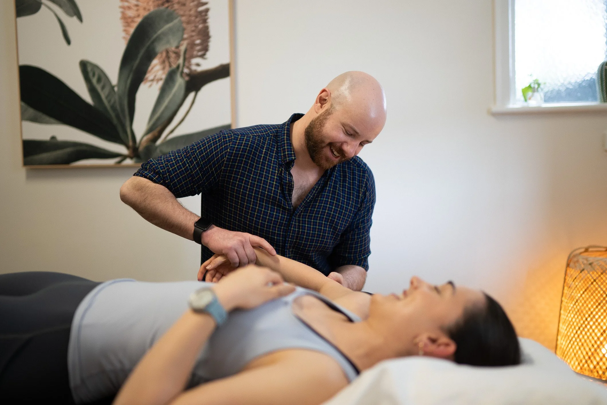 A man smiling and holding the arm of a woman lying in bed. The woman appears relaxed and is looking at the man. The scene takes place in a bedroom with a plant and artwork in the background.
