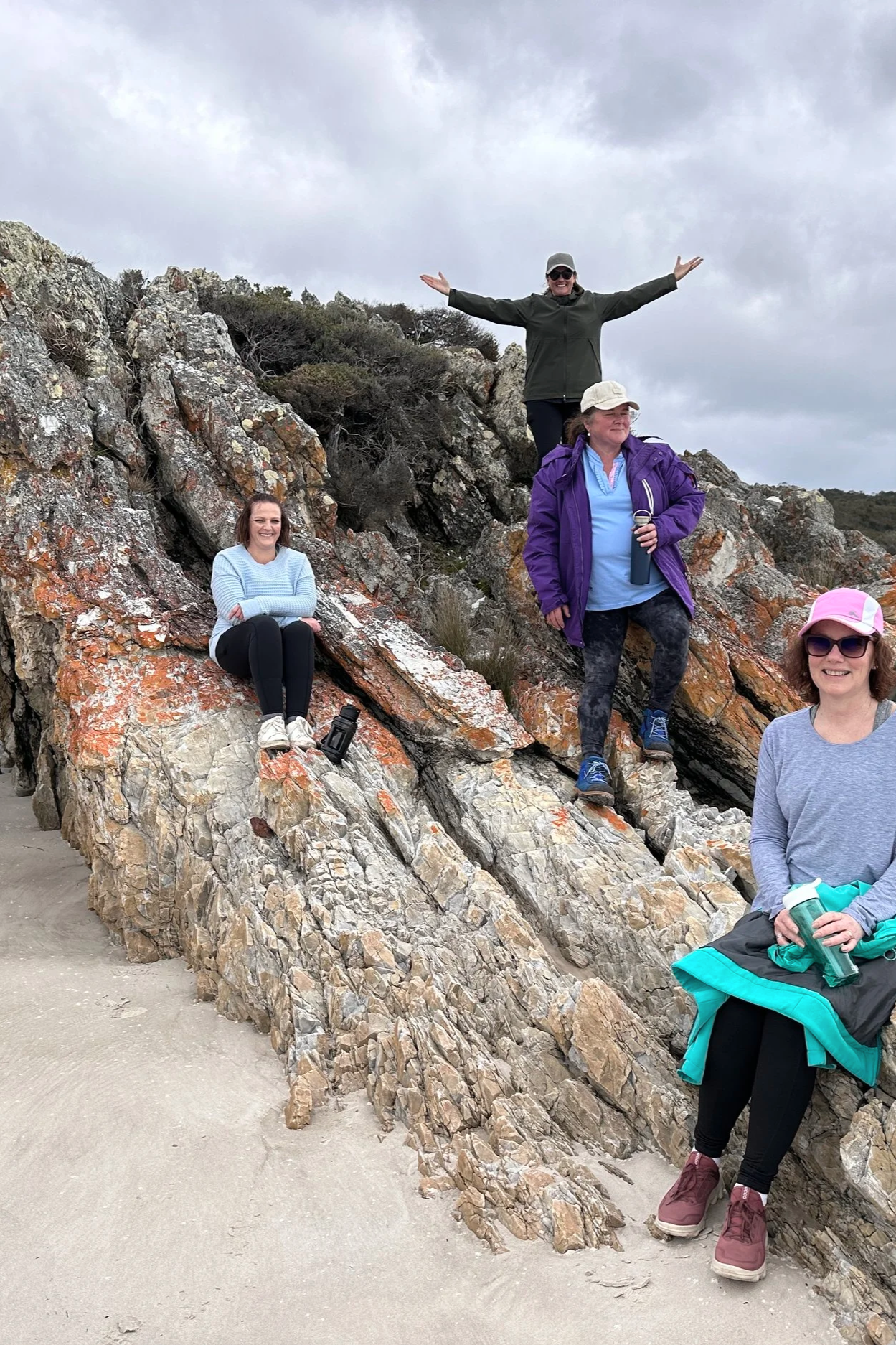 A group of five women enjoying a hike on a rocky coastal area. One woman is sitting on a large rock, others are standing or sitting on the rocks, dressed in outdoor clothing. The sky is overcast with clouds.