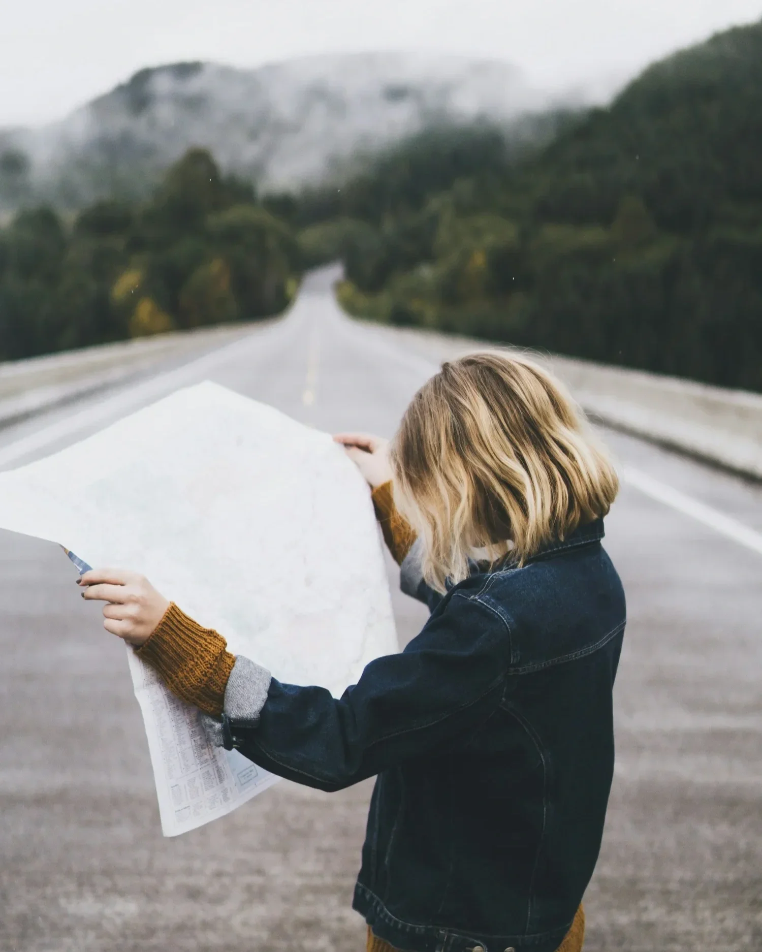 A young girl with blonde hair holding a map on an open road with mountains in the background.