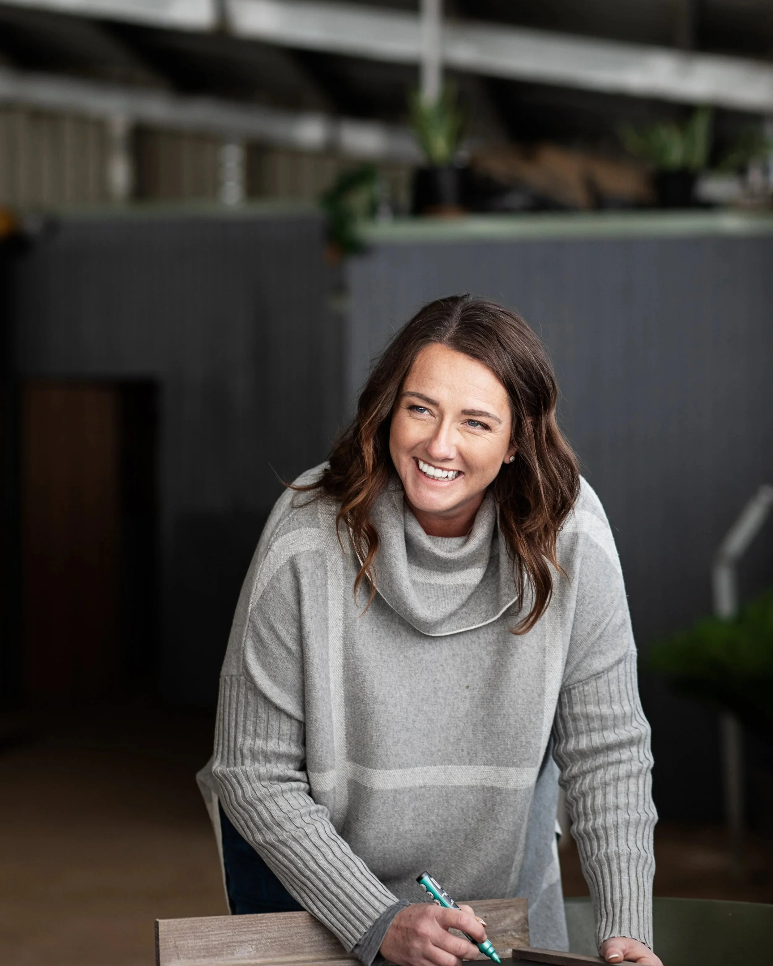 A woman with brown hair smiling, wearing a grey turtleneck sweater, signing a book with a teal pen.