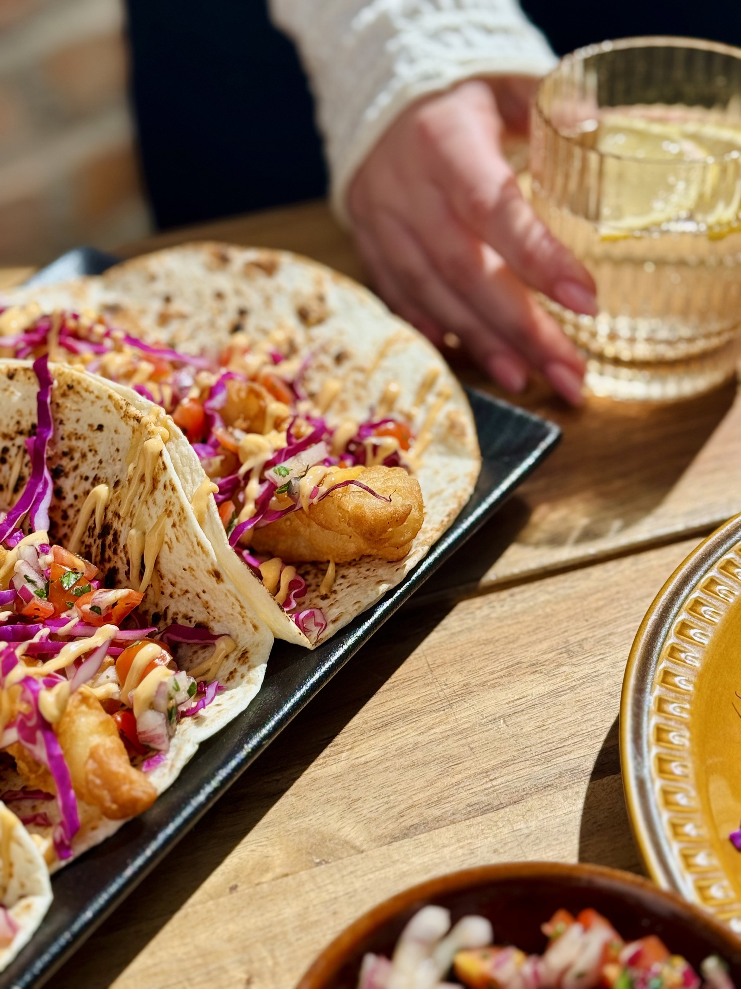 Close-up of a black rectangular platter with three tacos filled with colorful vegetables, shredded cabbage, served on a wooden table, with a glass bowl of yellow sauce and a gold-rimmed dish visible in the background.
