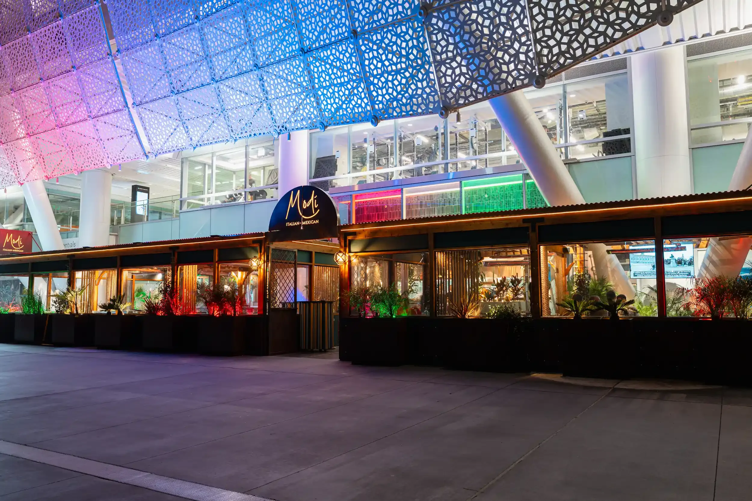 Night view of a modern shopping mall with colorful lighting, a restaurant named 'Moti,' and decorative illuminated panels on the building exterior.