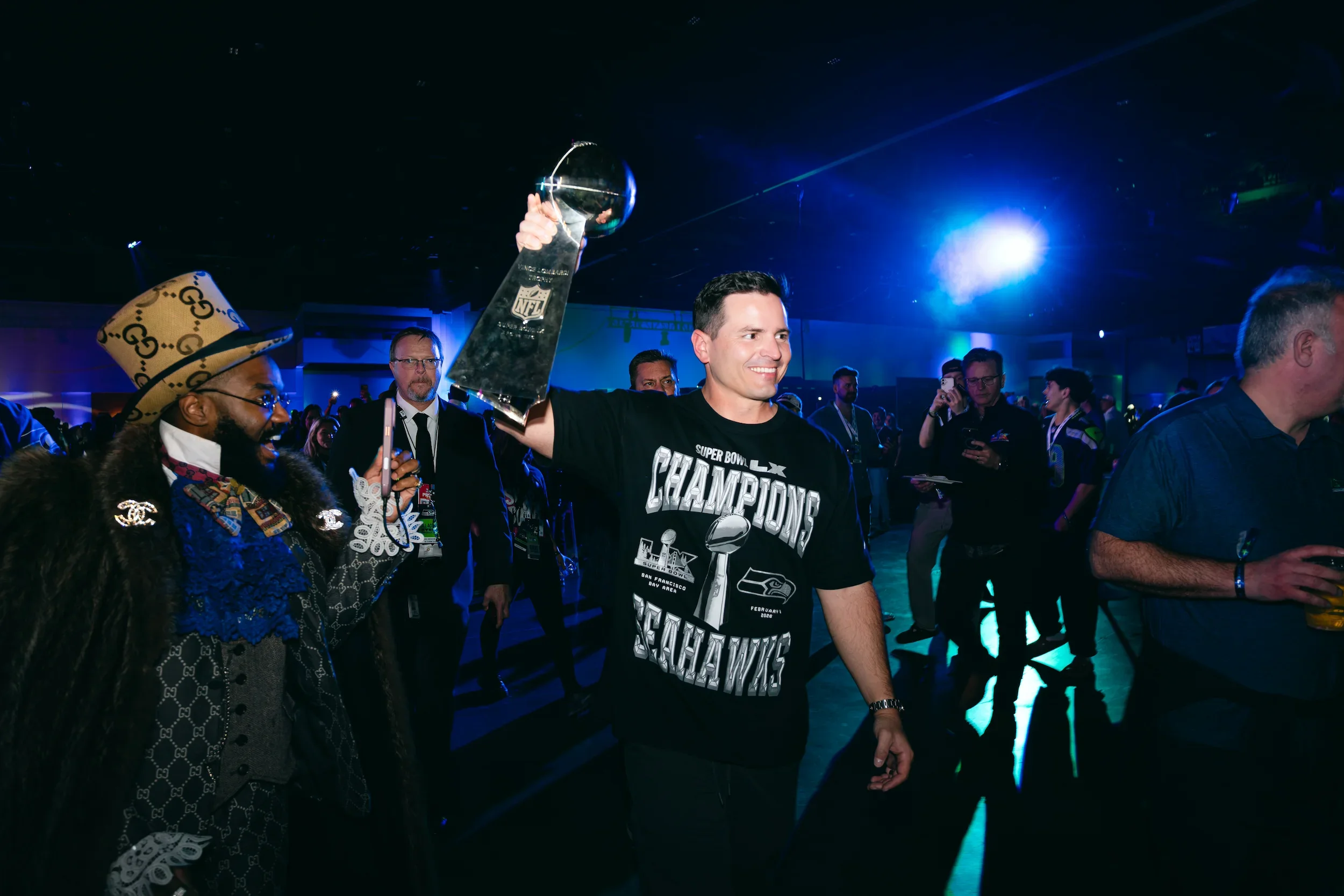 Mike Macdonald, wearing a black Seattle Seahawks Super Bowl Champions t-shirt, holding an NFL trophy at a celebration event, surrounded by other people.