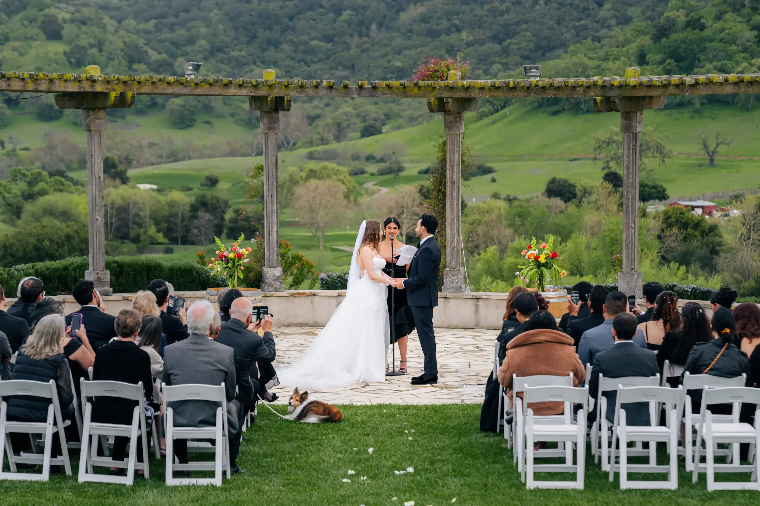 A wedding ceremony outdoors with a bride and groom holding hands, exchanging vows under a stone pergola. The officiant stands behind them with a microphone. Guests seated on white chairs watch the ceremony, some taking photos. A dog sits on the grass nearby. The background features lush green hills and trees.