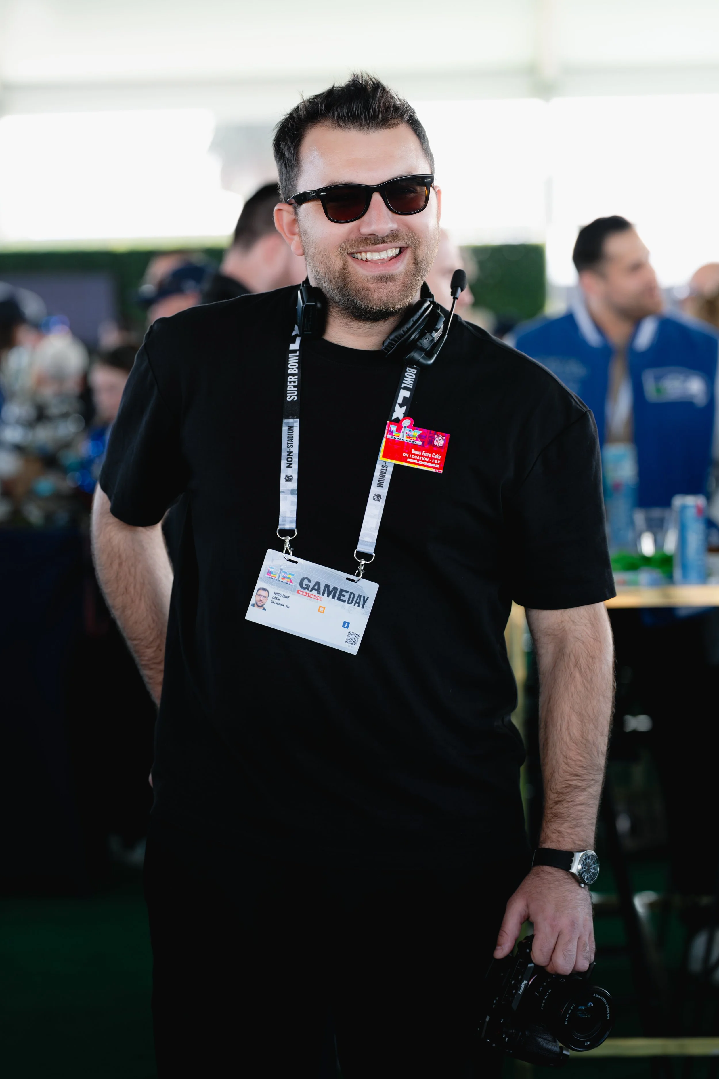 A man wearing sunglasses, a black t-shirt with a media pass and badge, holding a camera, and standing in an indoor area with other people.