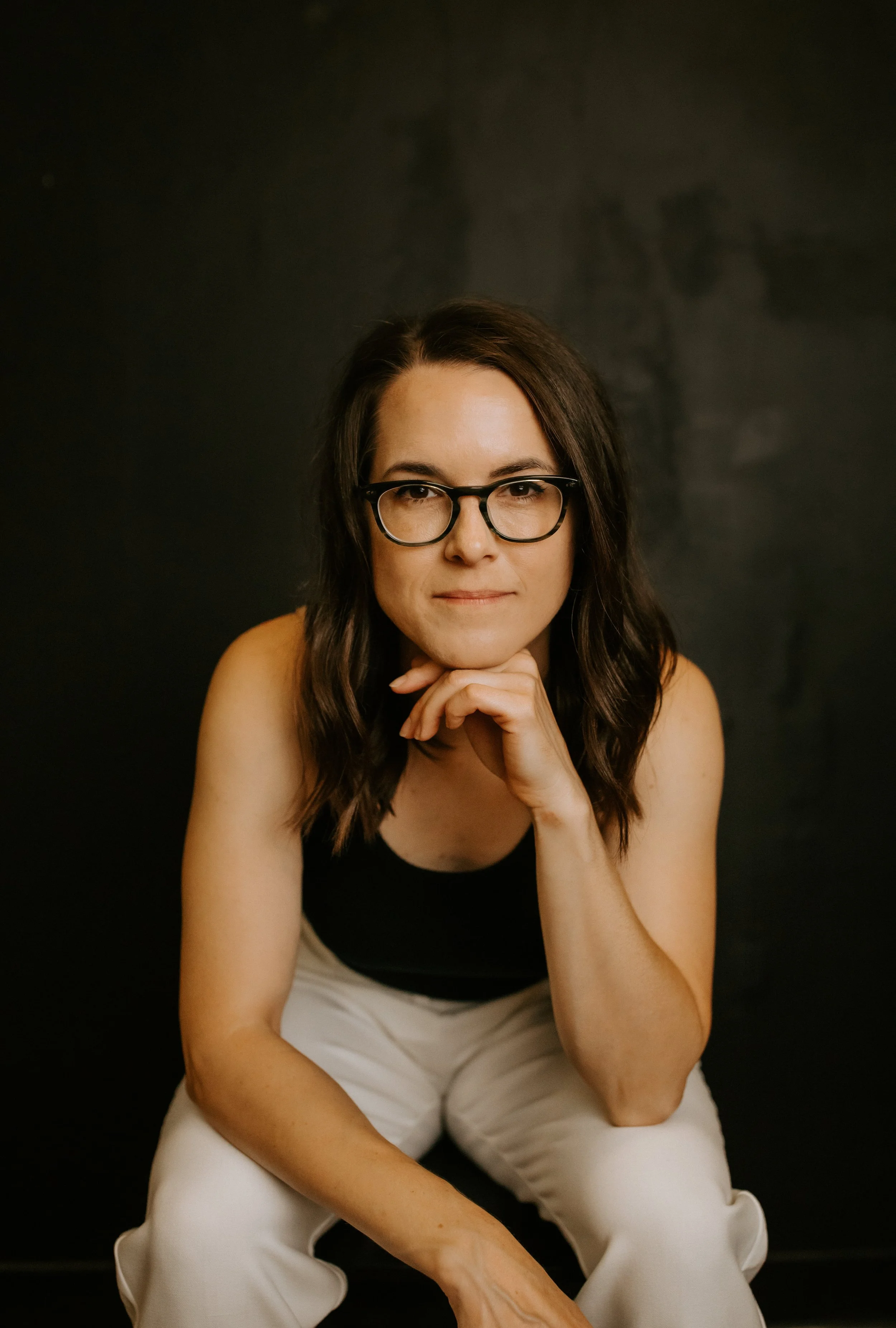 A woman with brown hair, glasses, and a black tank top sitting against a black background, resting her chin on her hand.