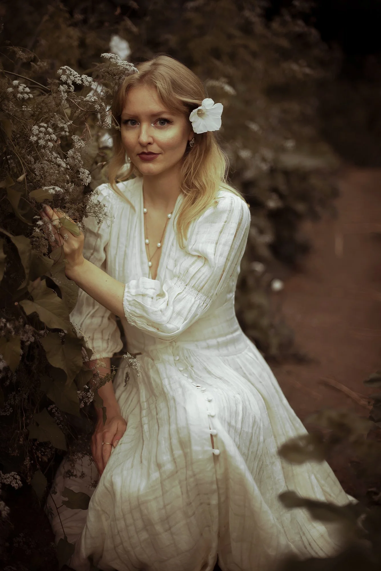 A woman with red hair, wearing a white dress with buttons, a pearl necklace, and an earring, sitting outdoors with brown foliage and plants around her. She has a large white flower tucked behind her ear.