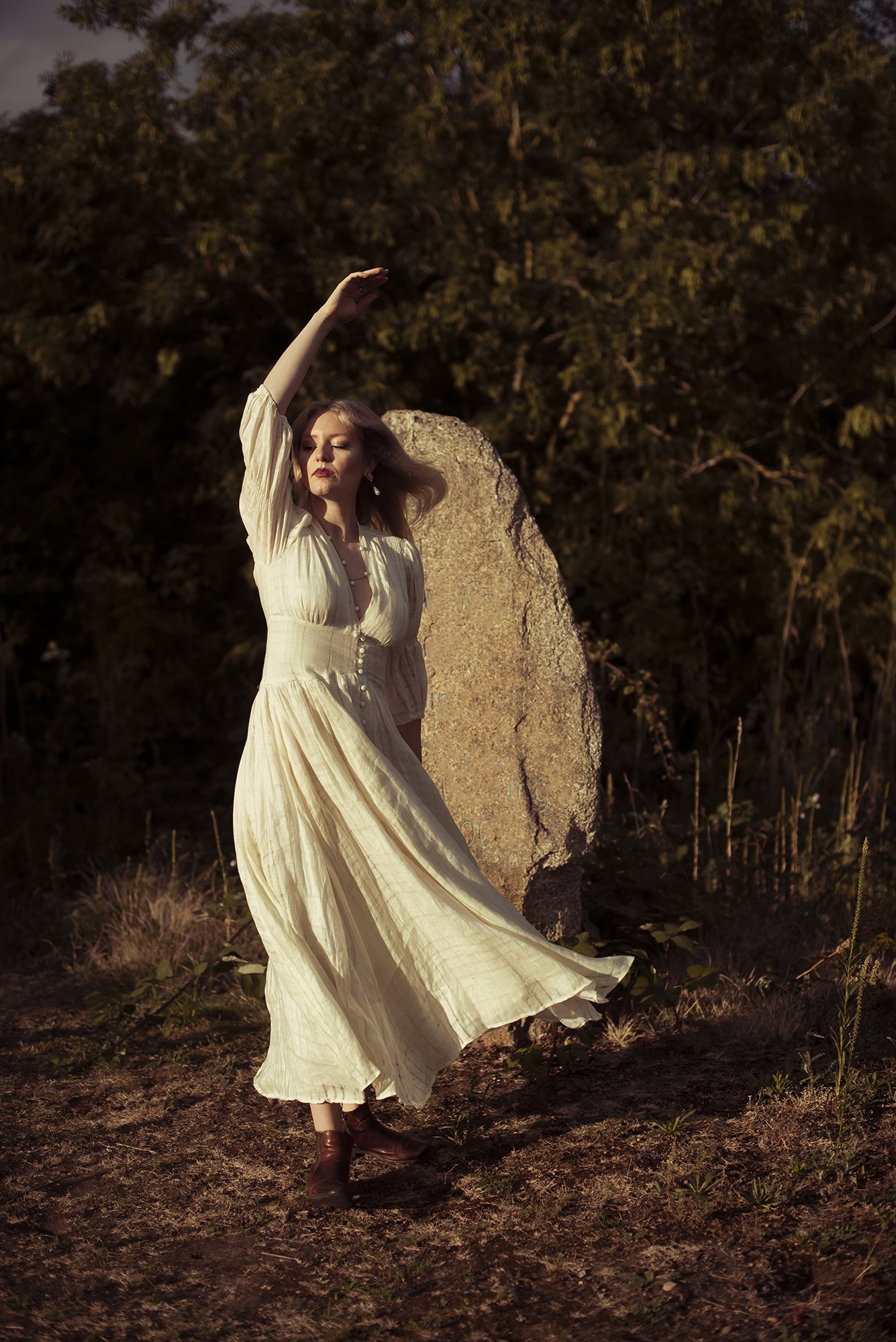 A woman with closed eyes in a flowing cream-colored dress stands outdoors at sunset, near a large rock and tall grass, with trees in the background.