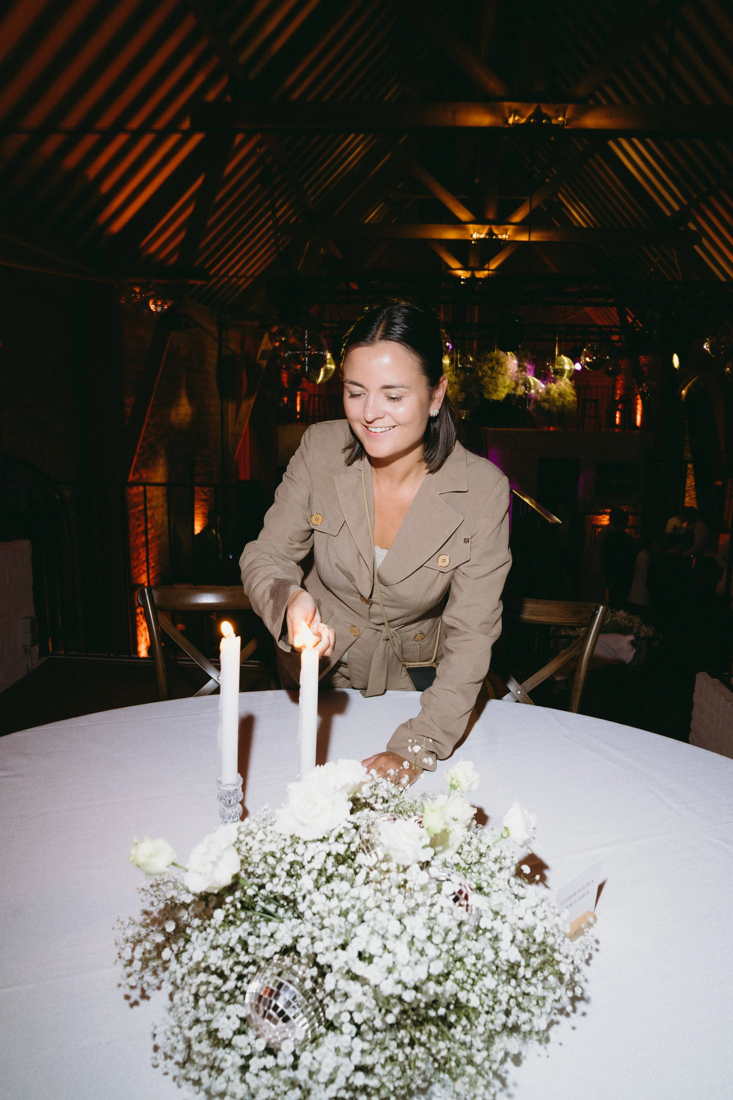 Vrouw die kaarsen aansteekt op een tafel met een witte tafelkleed en bloemendecoratie, in een sfeervolle ruimte met gedimd licht