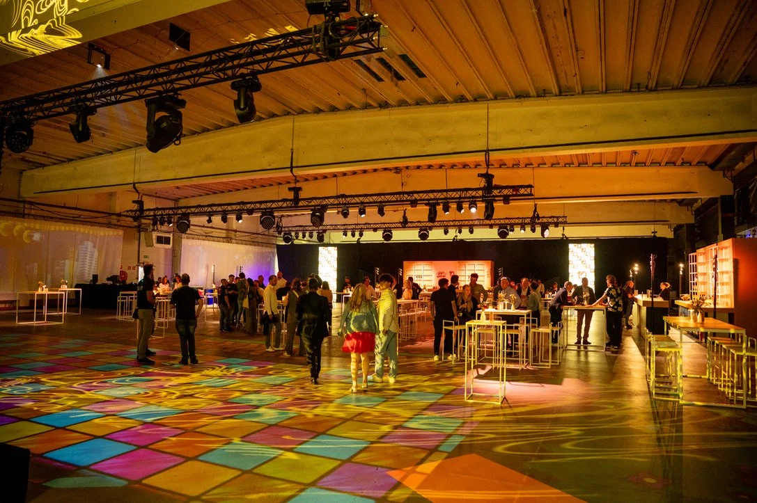 Een groot indoor evenement met mensen die rondlopen en socializen, verlicht met kleurrijke en zachte verlichting, met hoge plafonds en krachtig geluid- en lichtapparatuur.