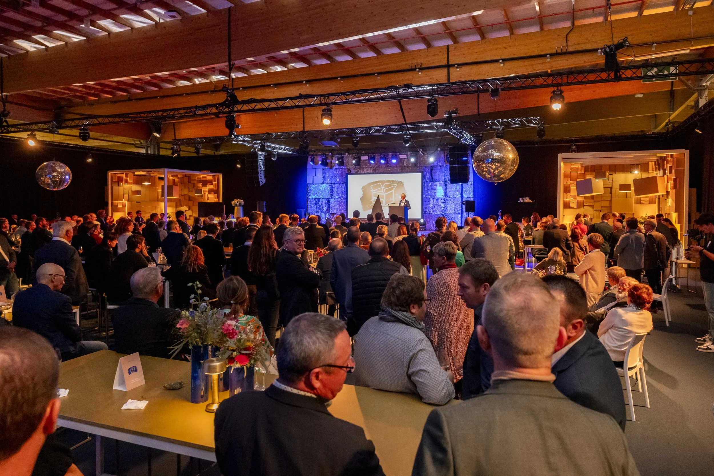 Grote groep mensen bij een evenement in een grote zaal, met een podium aan de achtergrond waar een spreker staat, decoratie met discoballen en warme verlichting.
