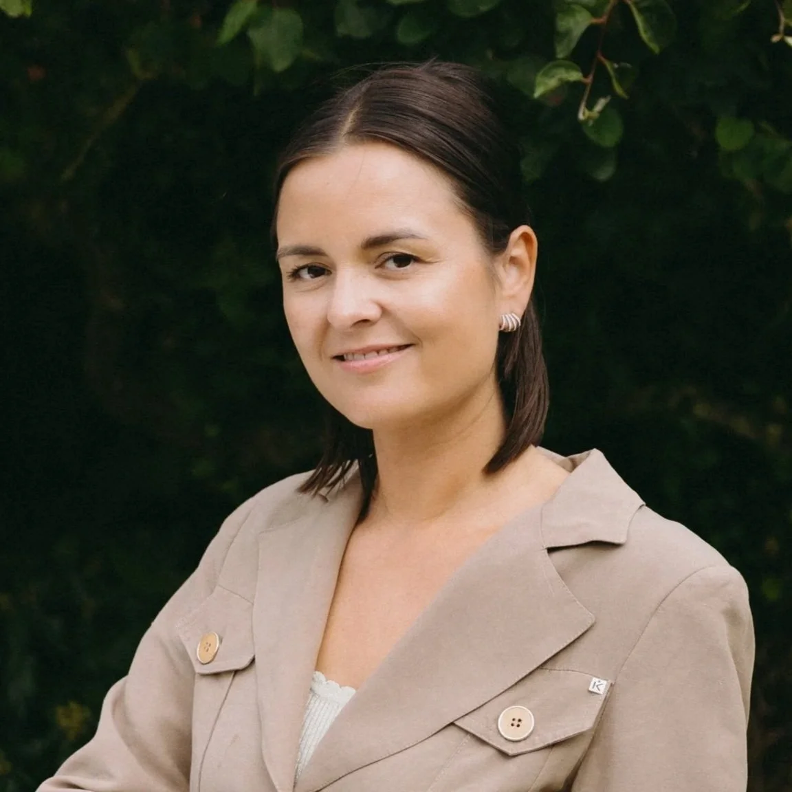 Portret van een vrouw met bruin, schouderlang haar, die in een beige jas staat voor een groene achtergrond.