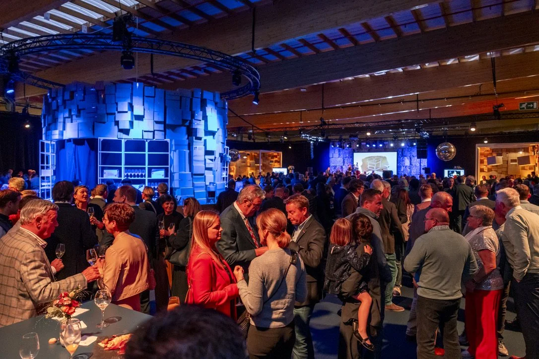 Grote groep mensen op een evenement of feestje in een zaal met blauwe en oranje verlichting, dicht op elkaar, met een groot scherm en decoraties op de achtergrond.