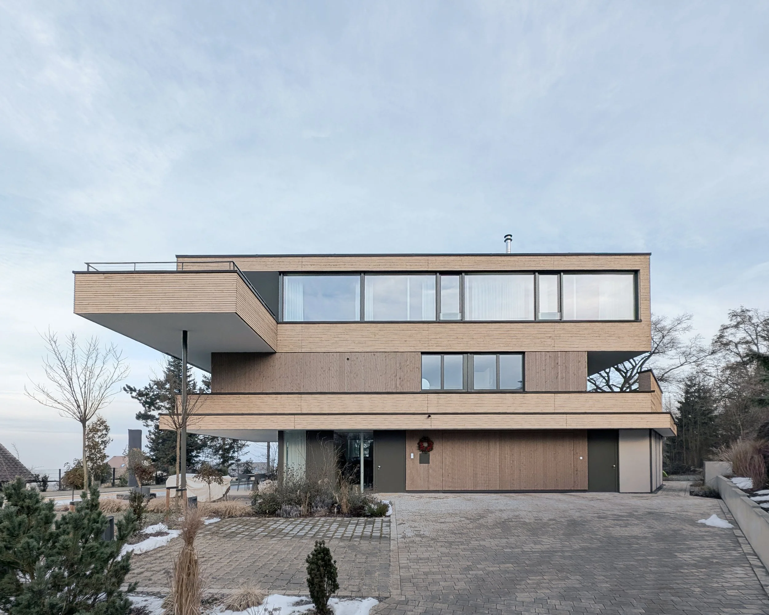 Modernes Haus mit mehreren Ebenen aus Holz und großen Fenstern, im Winter mit einigen Schneeflächen im Garten