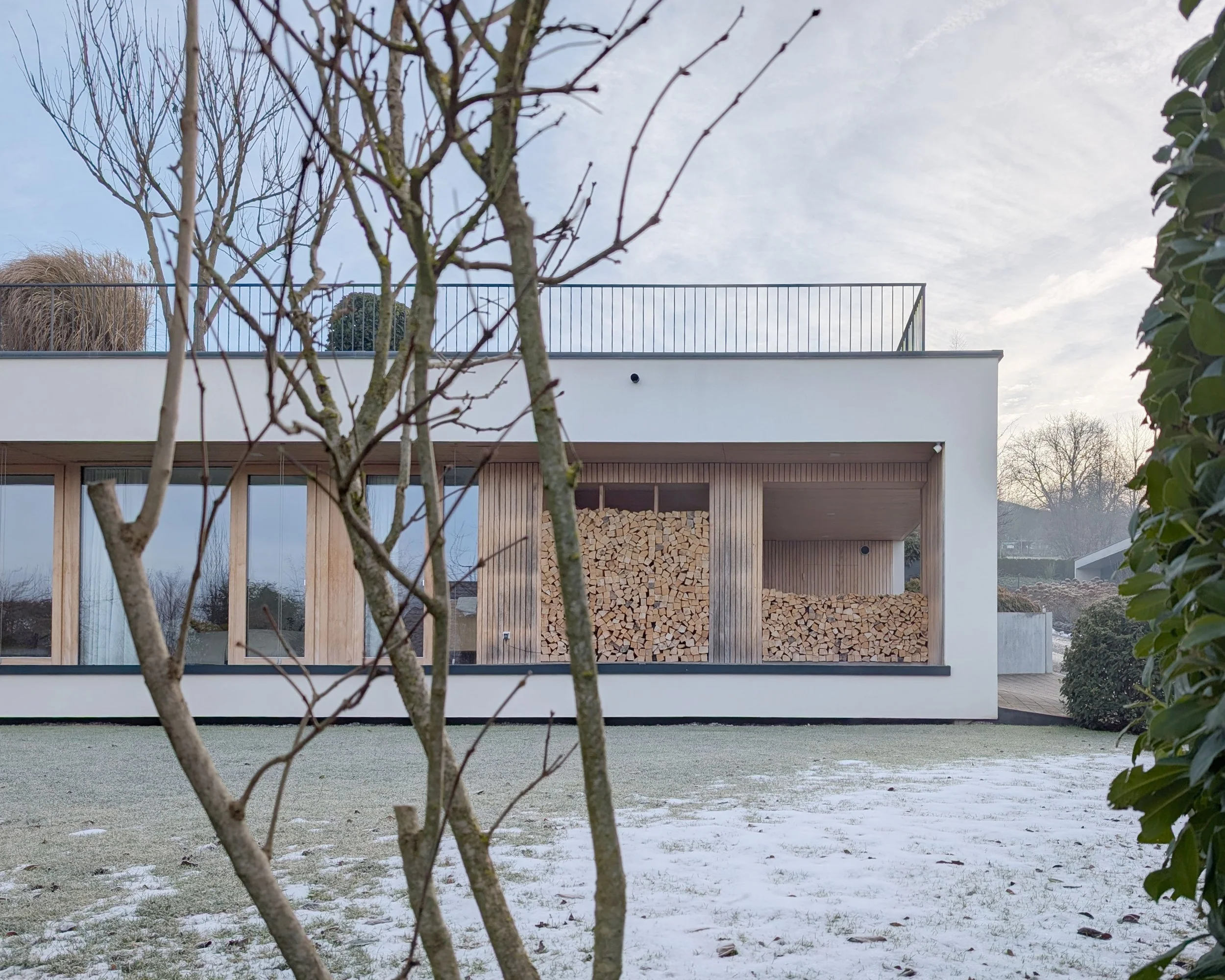 Moderner, weißer Haus mit Holzverkleidung und Holzstapel im Sichtbereich, umgeben von einem Garten mit niedrigem Gras und Bäumen, bei leicht frostigem Winterwetter, unter bewölktem Himmel.