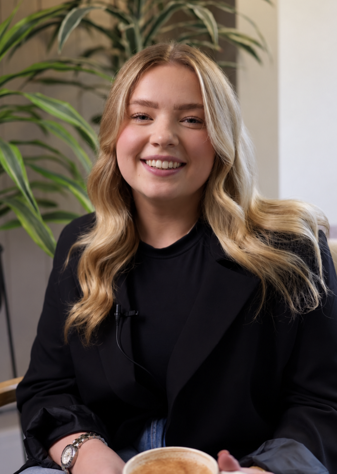 A young woman with long blonde hair and blue eyes sitting with her chin resting on her hand, looking thoughtfully to the side. She is wearing a black shirt and appears to be in a professional or waiting area. In the blurred background, a woman with dark hair is seen using a laptop.
