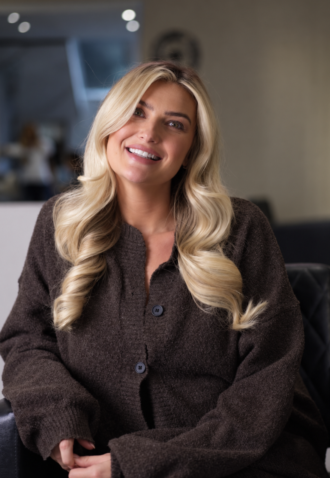 A woman with long blonde hair styled in loose curls, smiling and sitting in an indoor setting, wearing a dark brown button-up cardigan.