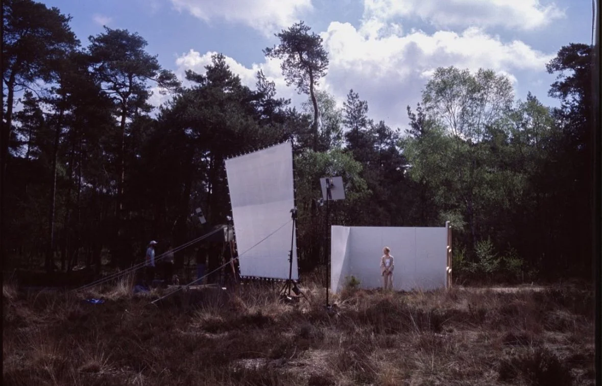A person standing in front of a white backdrop outdoors, with lighting equipment and crew nearby, set up for a photoshoot or filming in a natural, wooded area under partly cloudy skies.