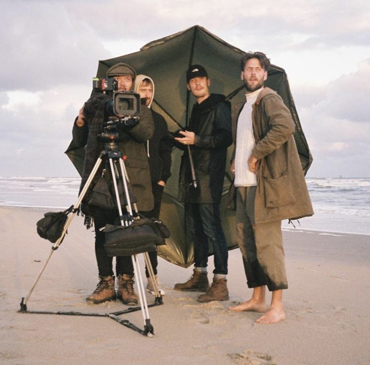 A film crew standing on the beach with a camera on a tripod, filming under a large umbrella with the ocean in the background.