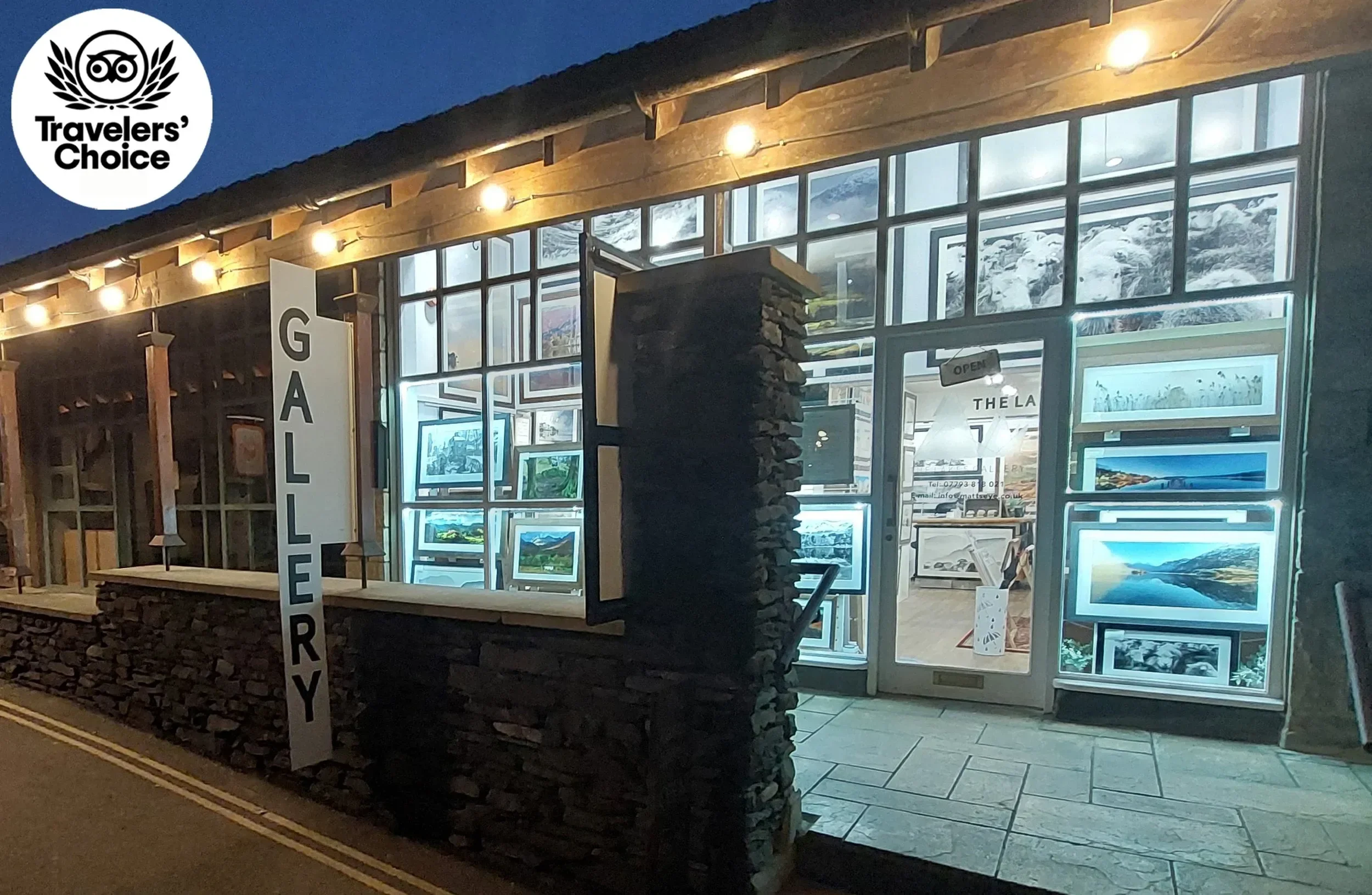 The exterior of an art gallery at night with a sign reading 'GALLERY' and framed landscape photographs displayed in the window, illuminated by exterior lights, and snow-capped mountains visible in the background.