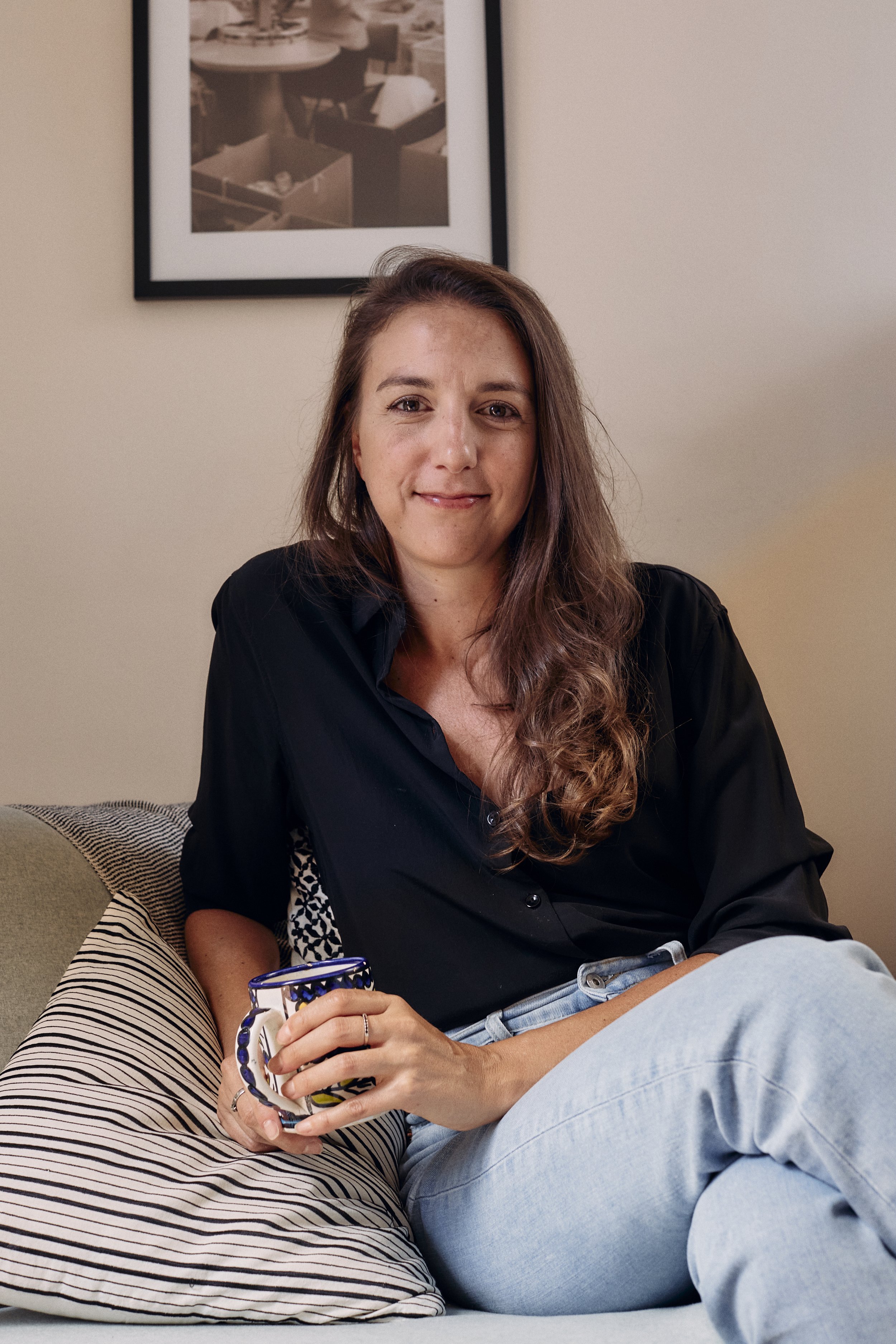Woman with long hair sitting on a couch holding a mug, wearing a black shirt and jeans, with a framed photo on the wall behind her.