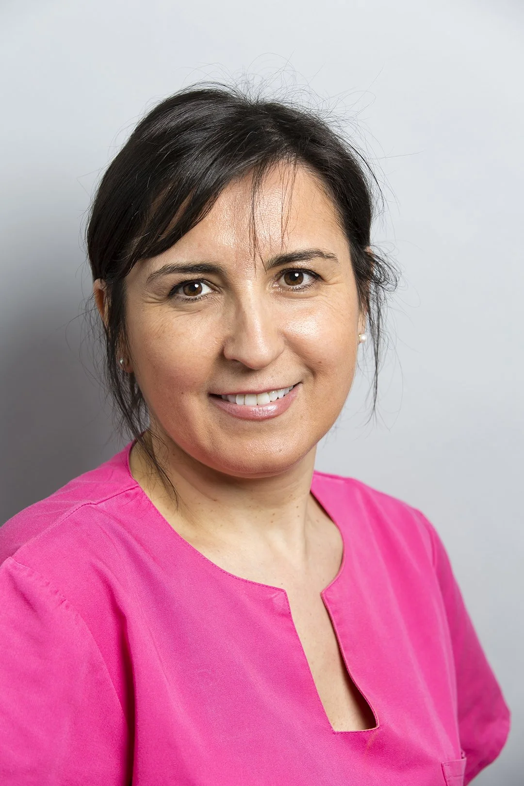 Une femme souriante portant une blouse rose sur fond gris clair.