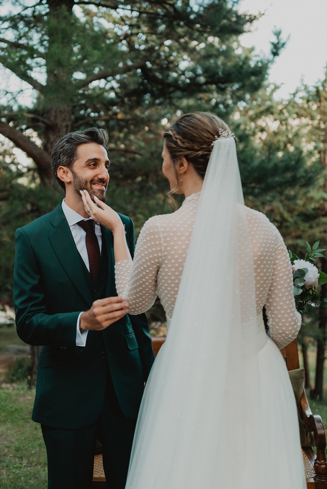 Una pareja vestida de novios en un entorno natural. La novia lleva un vestido blanco con velo y el novio un traje oscuro. Están mirándose con cariño.