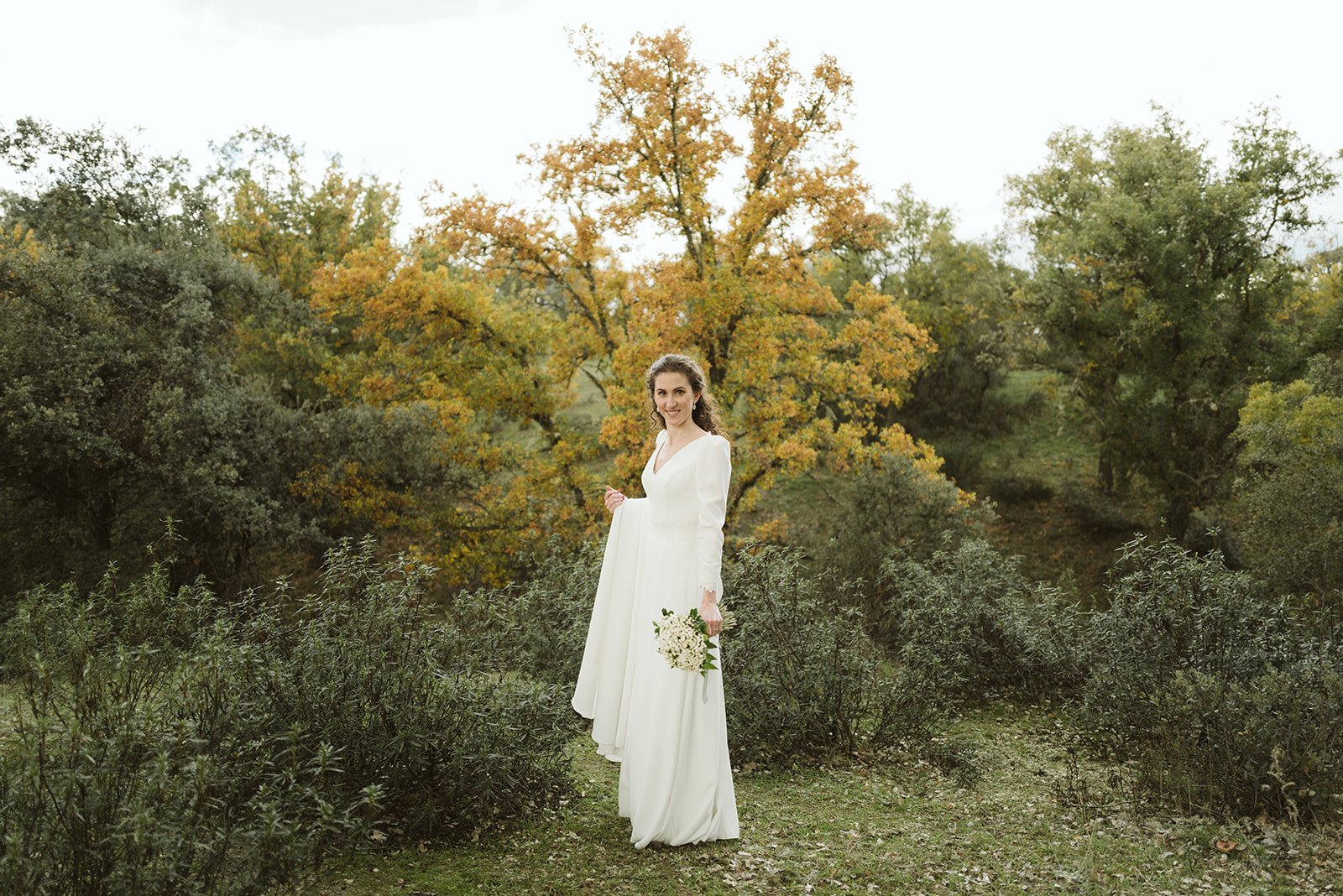 Mujer en vestido de novia blanco con ramo de flores, rodeada de naturaleza y árboles con hojas otoñales.