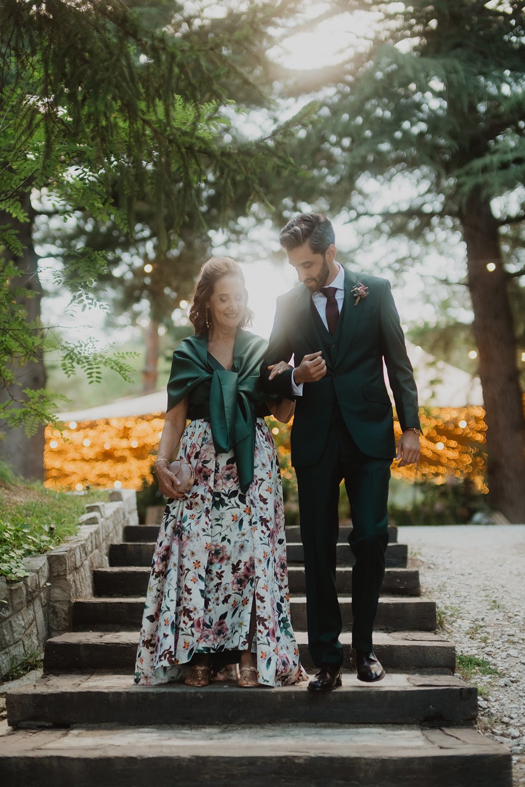Pareja elegante caminando por escaleras al aire libre, rodeados de árboles y luces decorativas.